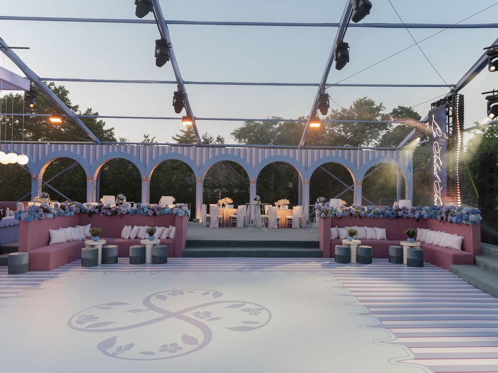 Elegant outdoor event space with pink and white seating, floral decorations, and a long table set up for a formal gathering, framed by arched structures and string lights at dusk.