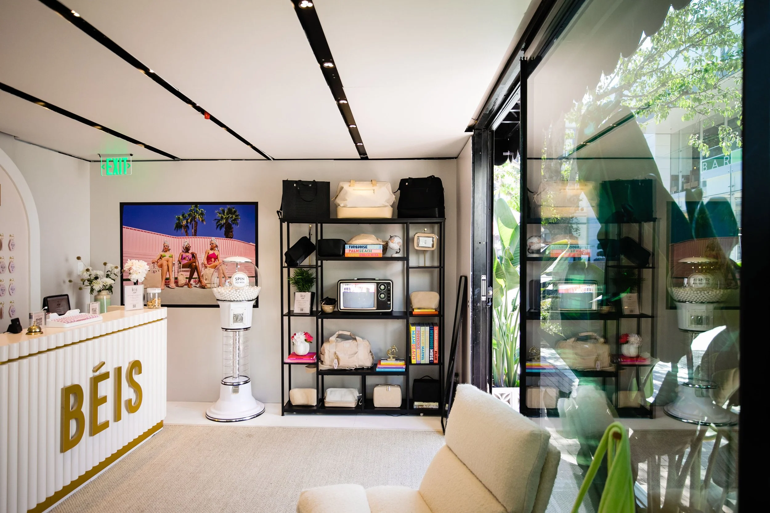 Interior view of a gift shop with a white counter, gold 'BÉIS' lettering, black shelving unit with bags, books, and decor, a large framed photo of three women sitting outdoors, an exit sign, a vacuum-shaped golf ball dispenser, a small beige sofa, an
