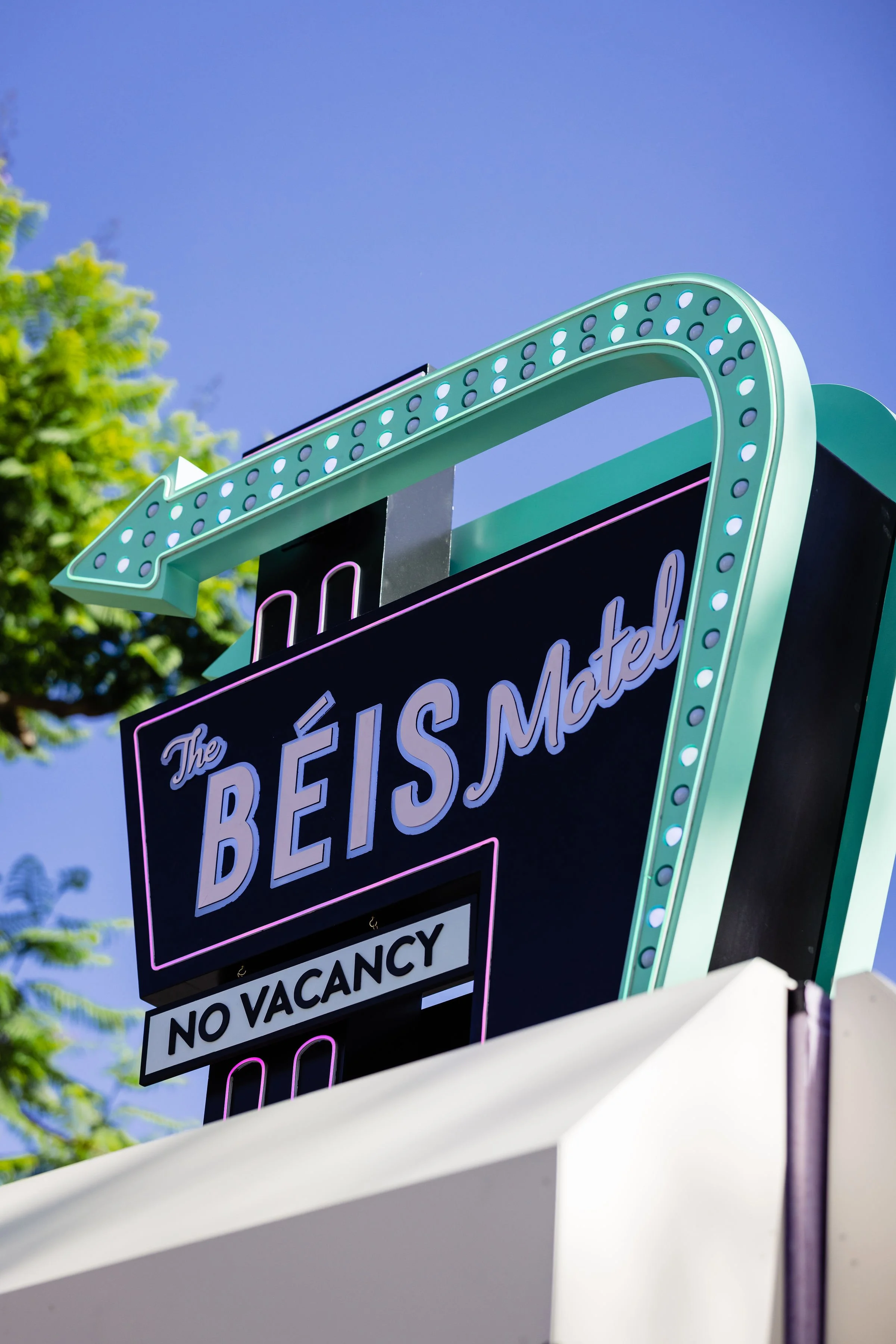 Neon sign for The BEIS Motel, status of No Vacancy, under a blue sky with green trees in the background.
