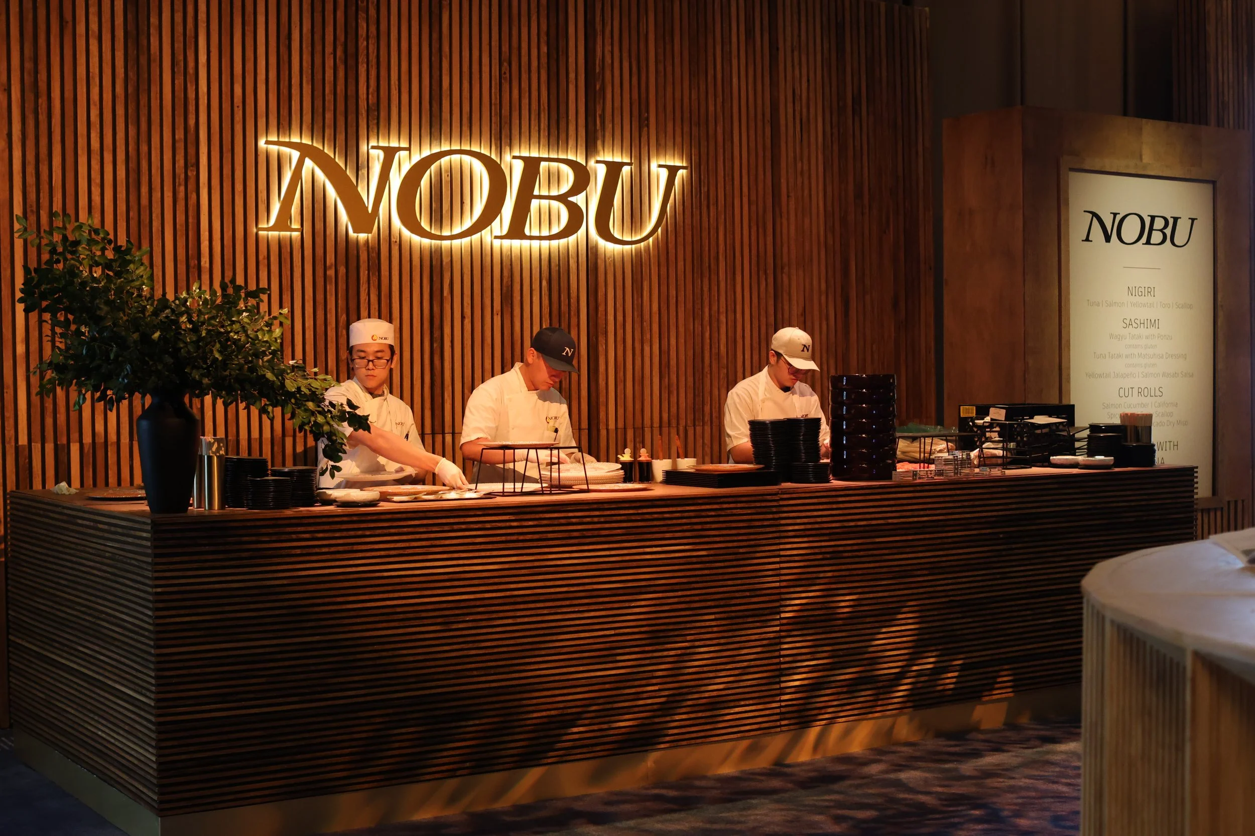 Three sushi chefs behind a wooden counter with a large illuminated 'NOBU' sign on wooden wall, preparing food in a restaurant.