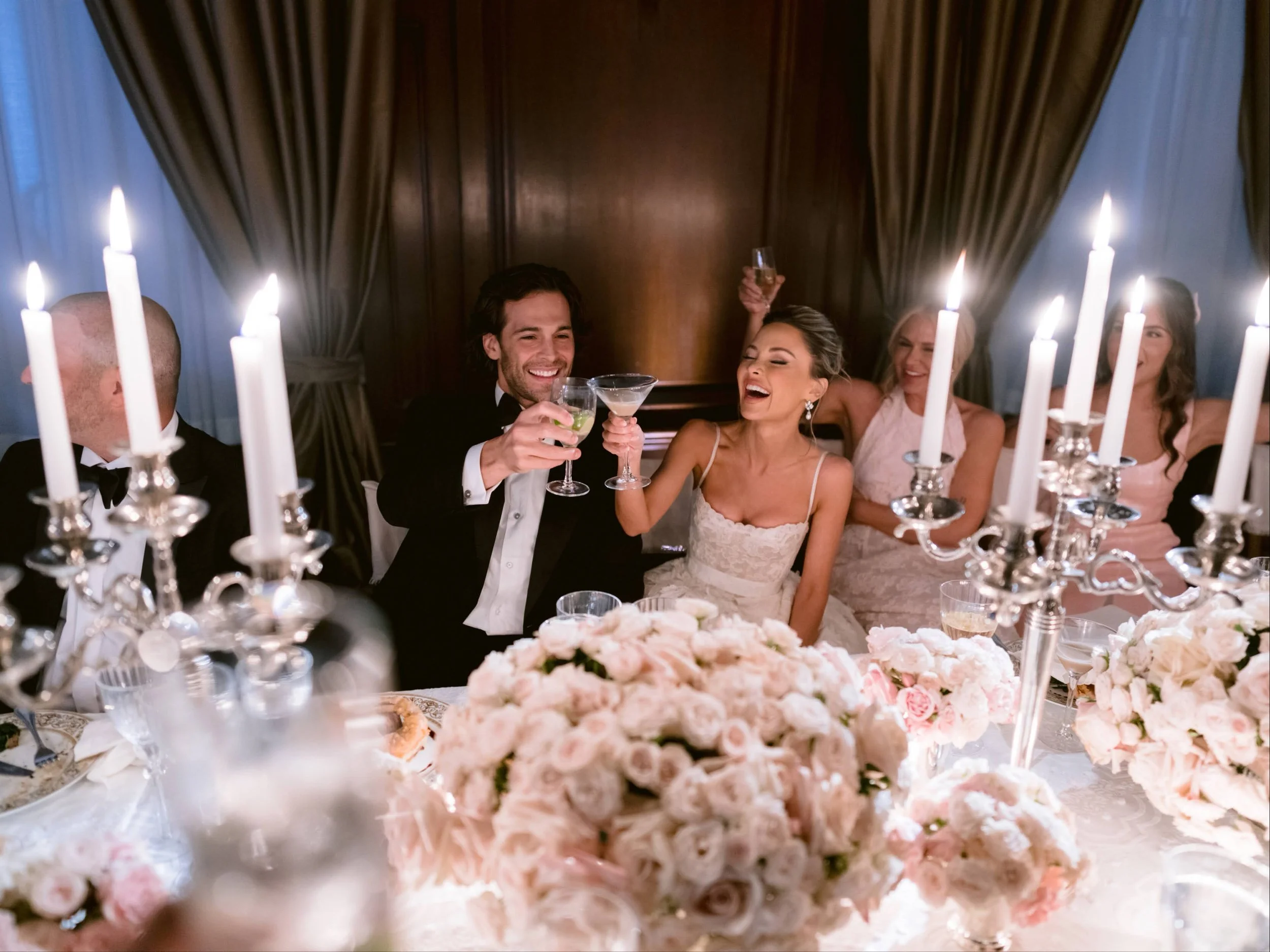 A wedding reception celebration with a bride, groom, and guests raising glasses for a toast, seated at a table adorned with pink and white floral arrangements and lit candles.