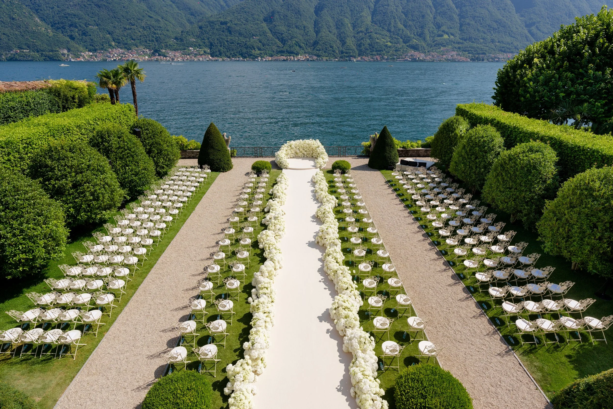 Outdoor wedding ceremony setup with white chairs arranged in rows on either side of a white aisle, decorated with white flowers, overlooking a lake with mountains in the background.