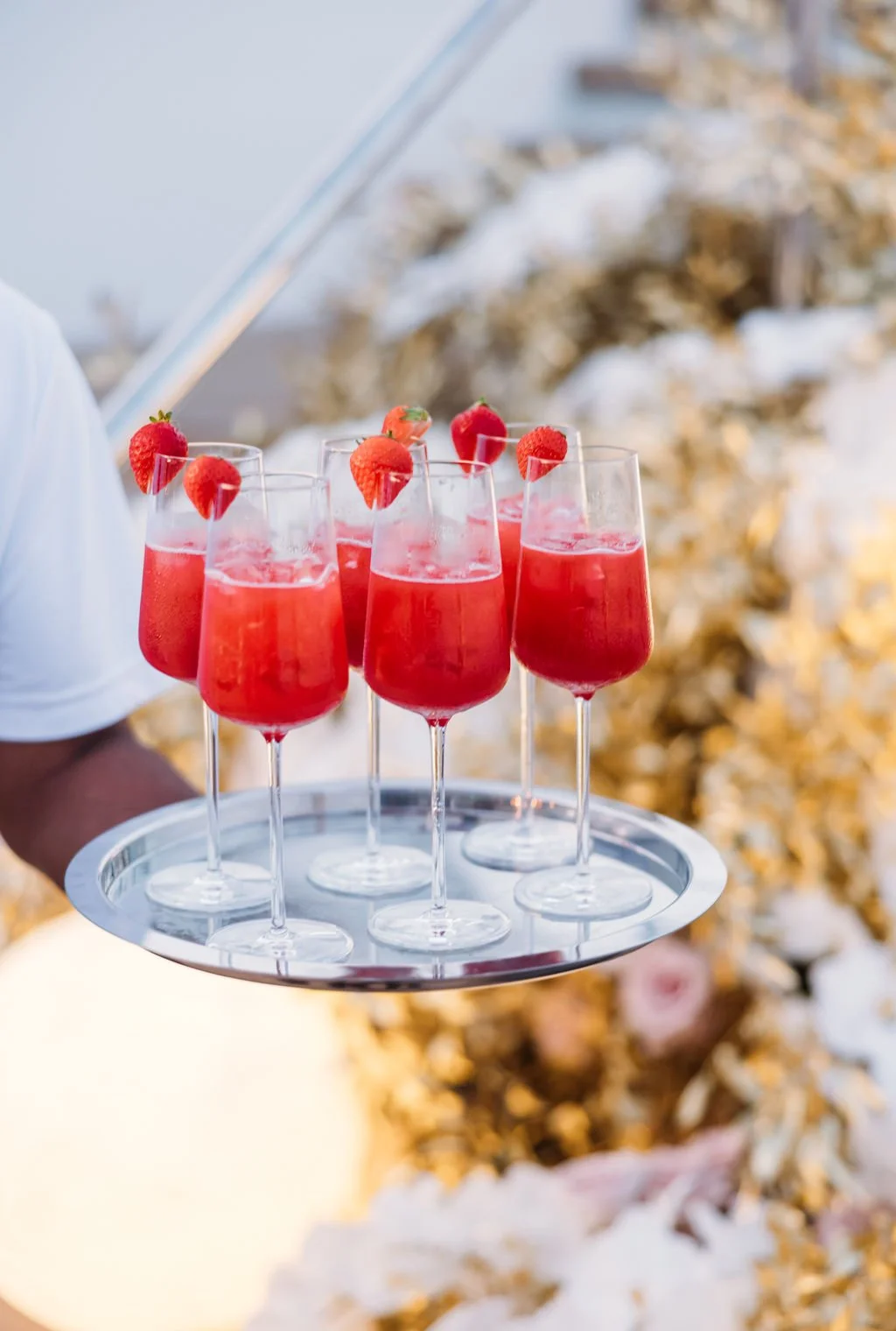 A tray of six glasses filled with red strawberry beverages, garnished with strawberries on top, held by a server at an event.