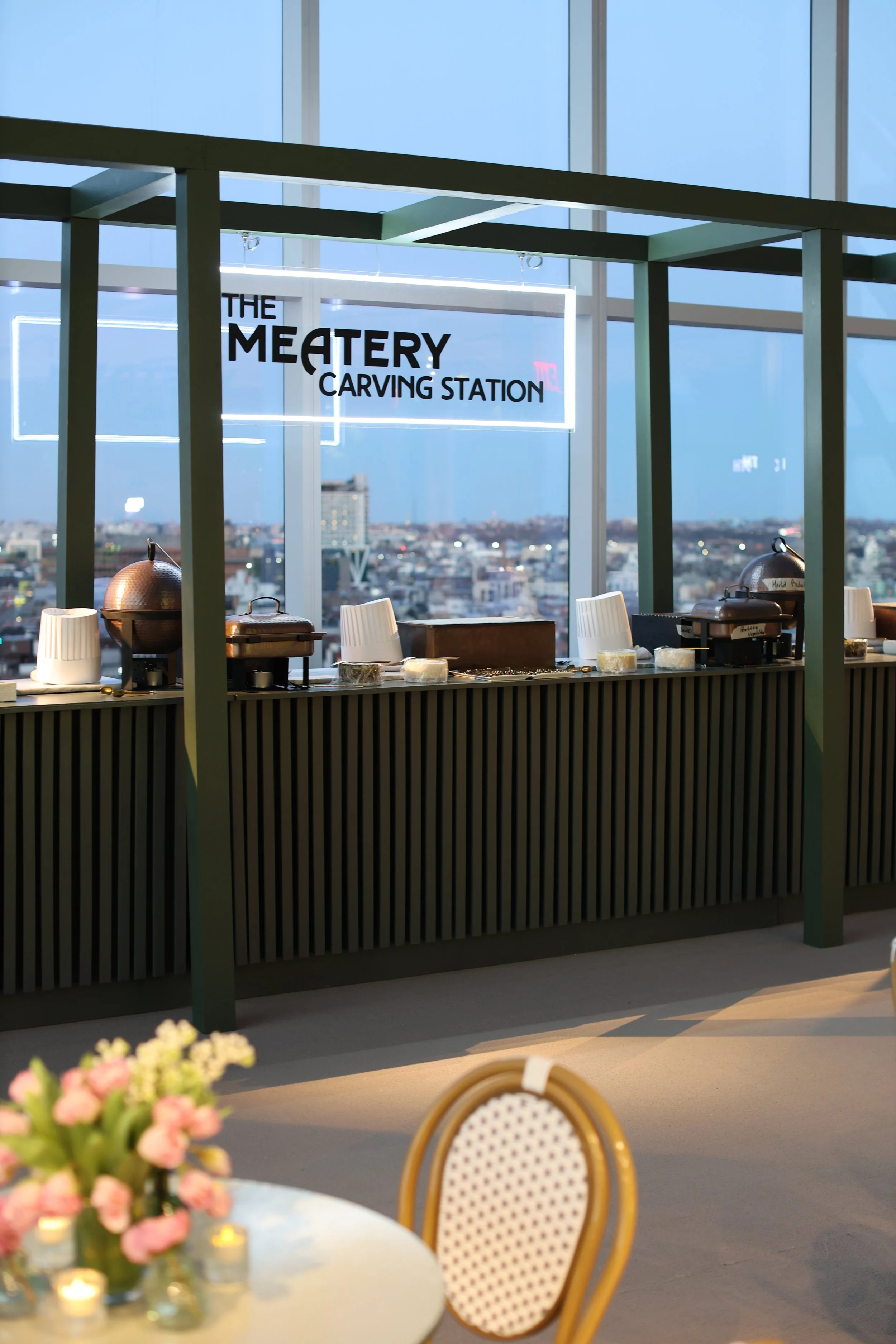 Interior of a modern event space or restaurant with a carving station setup, neon sign that reads 'The Meatery Carving Station,' and a city skyline visible through large windows in the background.