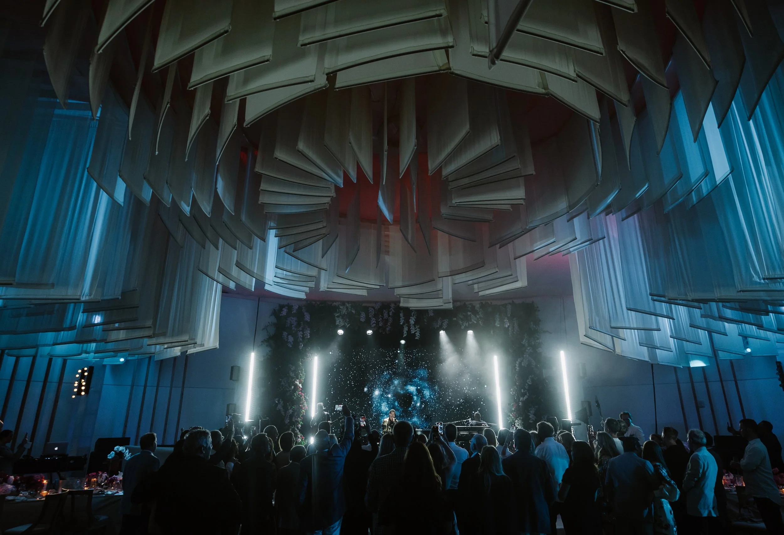 An indoor concert or event with a crowd of people in front of a stage featuring a digital cosmic display, with hanging book-like panels ceiling decoration and dramatic lighting.