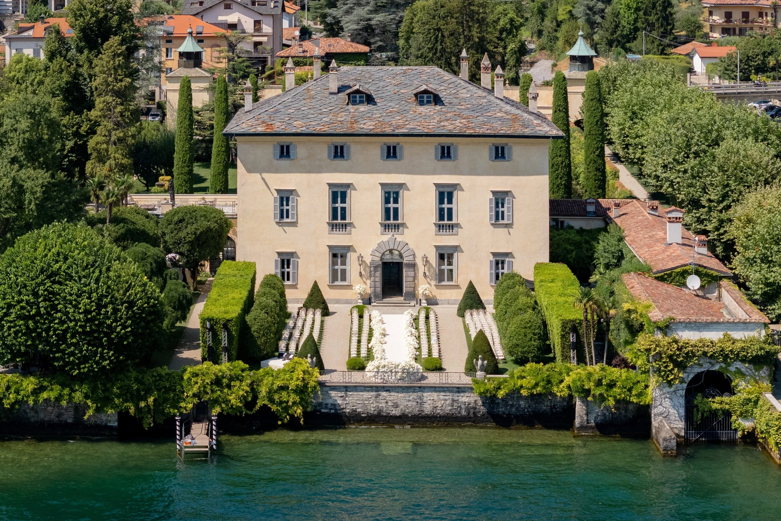 A large, elegant house situated on the waterfront, surrounded by lush greenery and landscaped gardens with a fountain and pathway leading to the entrance