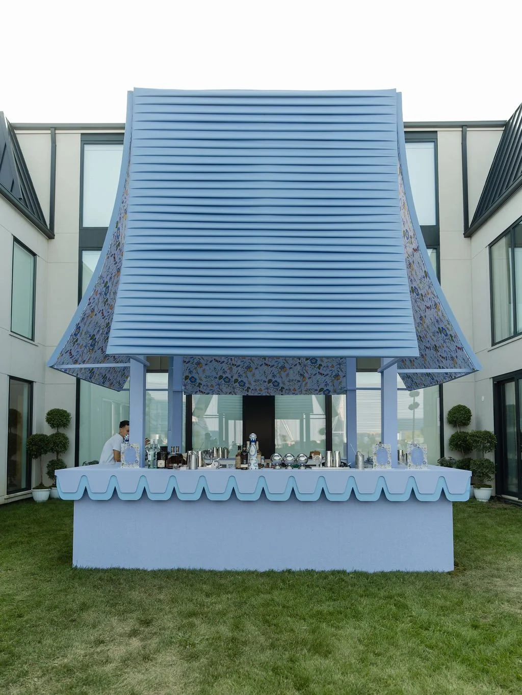 Outdoor bar setup with a large blue awning, white table, and various drink bottles, situated in a courtyard of modern buildings with potted plants.