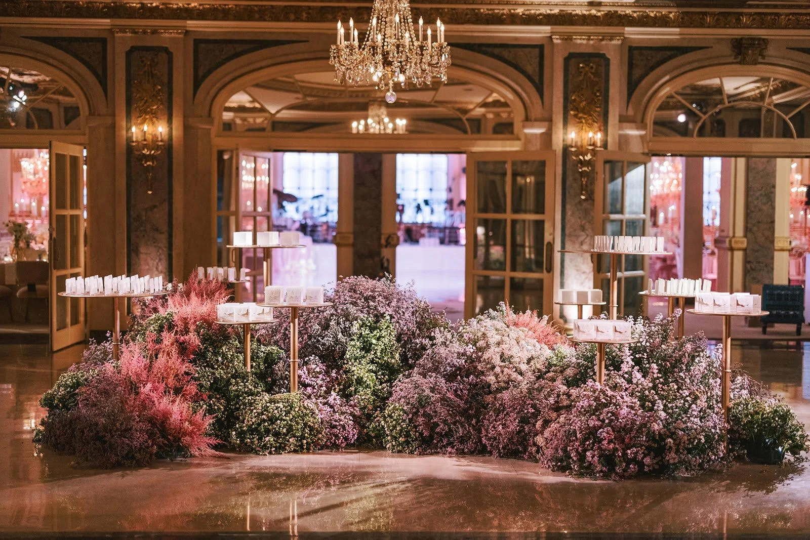 Elegant indoor setting with a grand chandelier, floral arrangements, candles, and multiple tables set for an event or celebration.