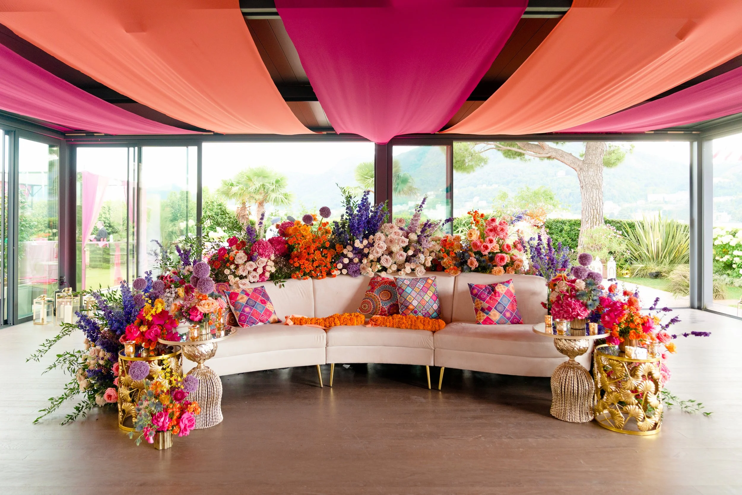 A floral wedding or event setup with a curved beige sofa decorated with colorful pillows, surrounded by an abundance of vibrant flowers, gold side tables, and pink and orange drapery on the ceiling, with large windows revealing a lush outdoor garden.