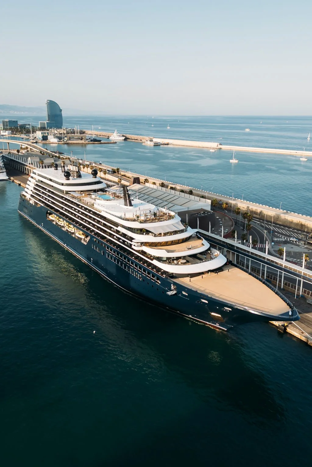 An aerial view of a cruise ship docked at a harbor, with a city skyline and the ocean in the background, including sailboats and the iconic W Barcelona hotel.