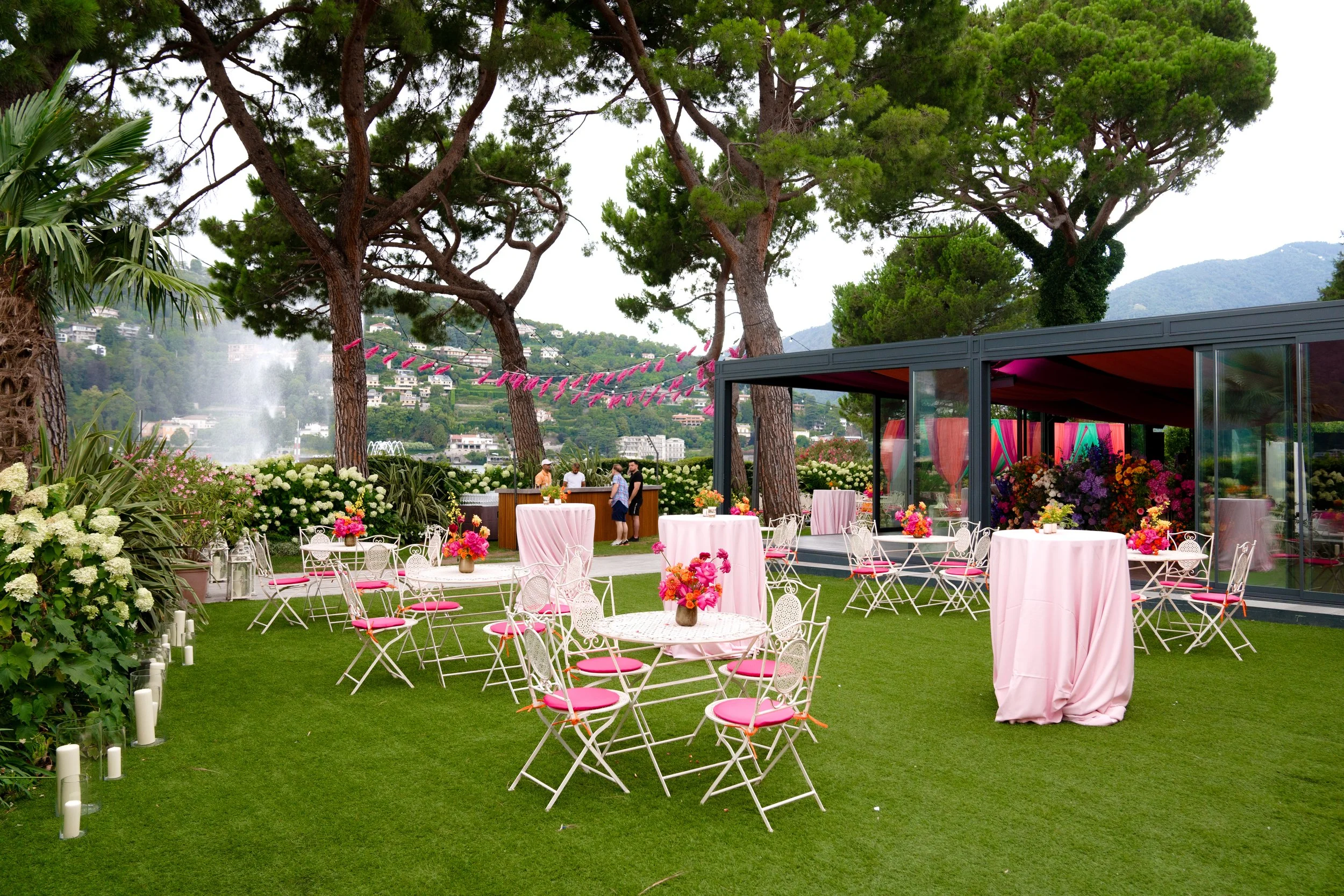 Outdoor event setup with pink tablecloths, floral centerpieces, and chairs on a grassy area surrounded by trees and flowers, with mountains and a fountain in the background.