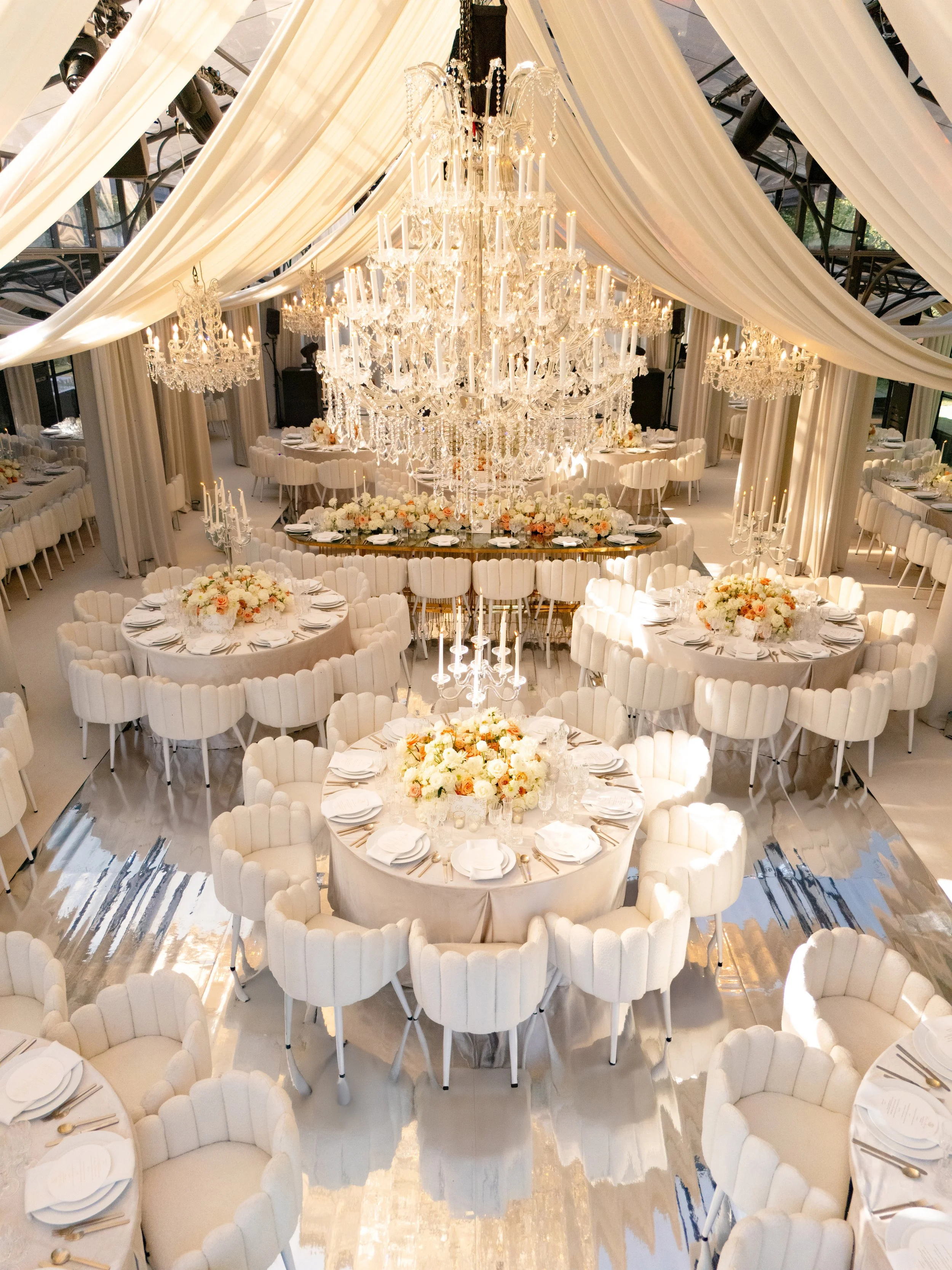 Elegant event space decorated with large chandelier, draped fabric ceiling, round tables with white chairs and floral centerpieces, and golden reflective flooring.