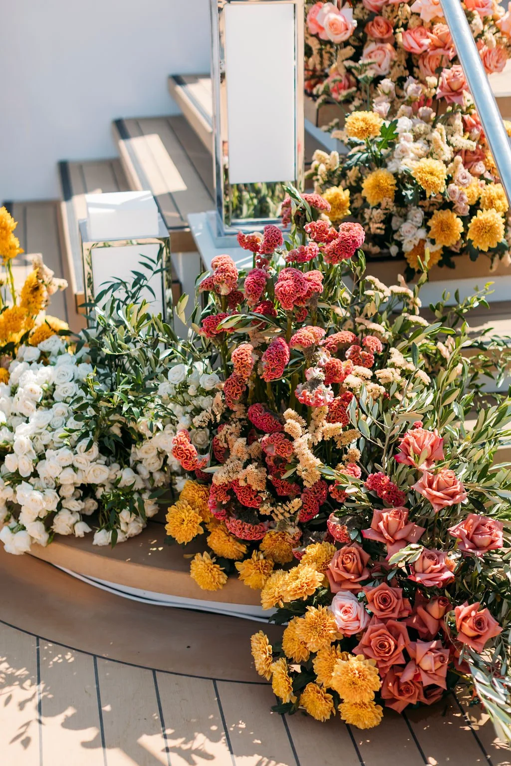 Colorful flowers, including roses, carnations, and chrysanthemums, arranged outdoors on a deck with sunlight.