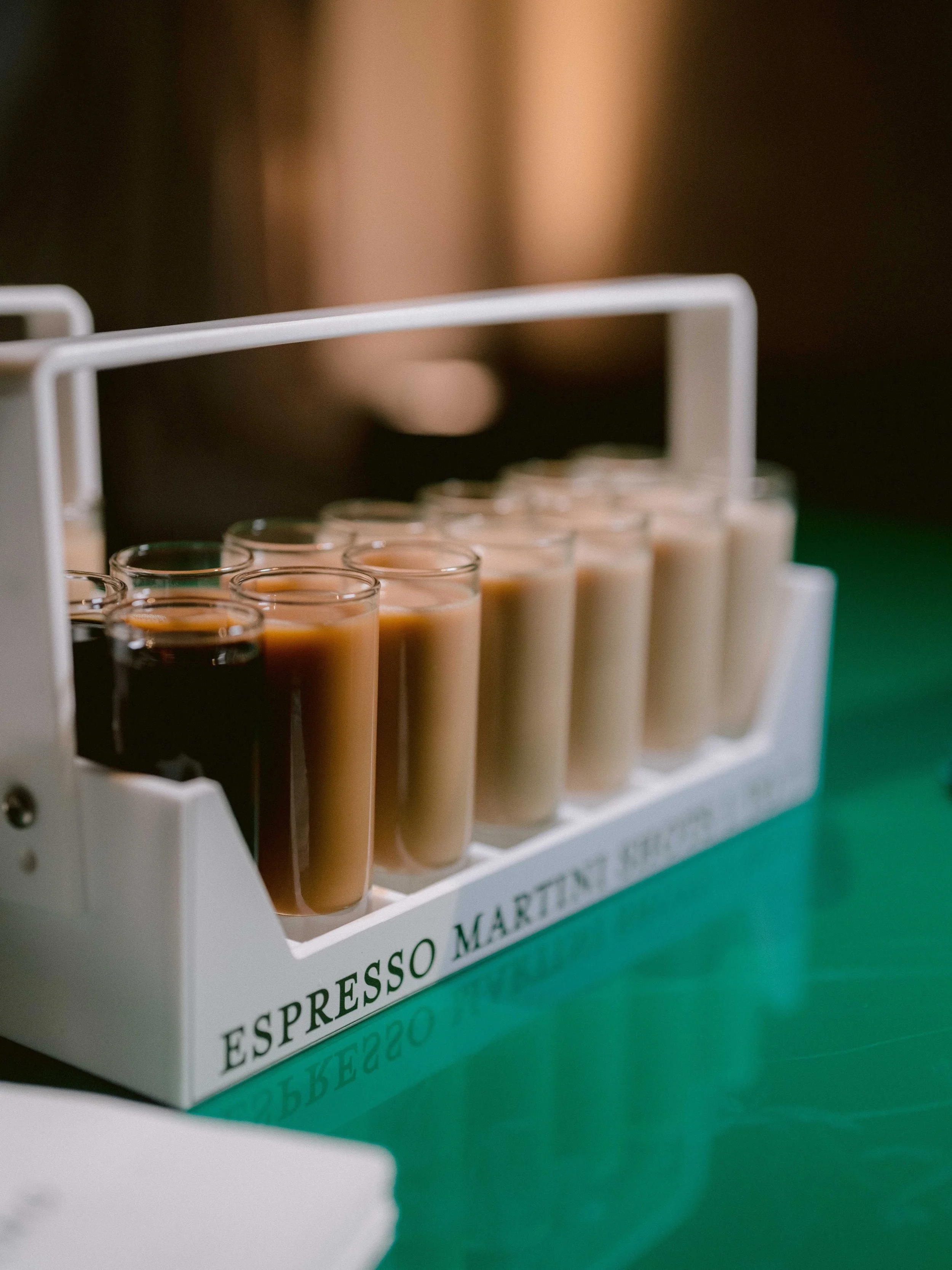 A white tray with various small glasses filled with different shades of coffee and tea, labeled with the words 'Espresso Martini & Tea'. The tray is on a green surface.