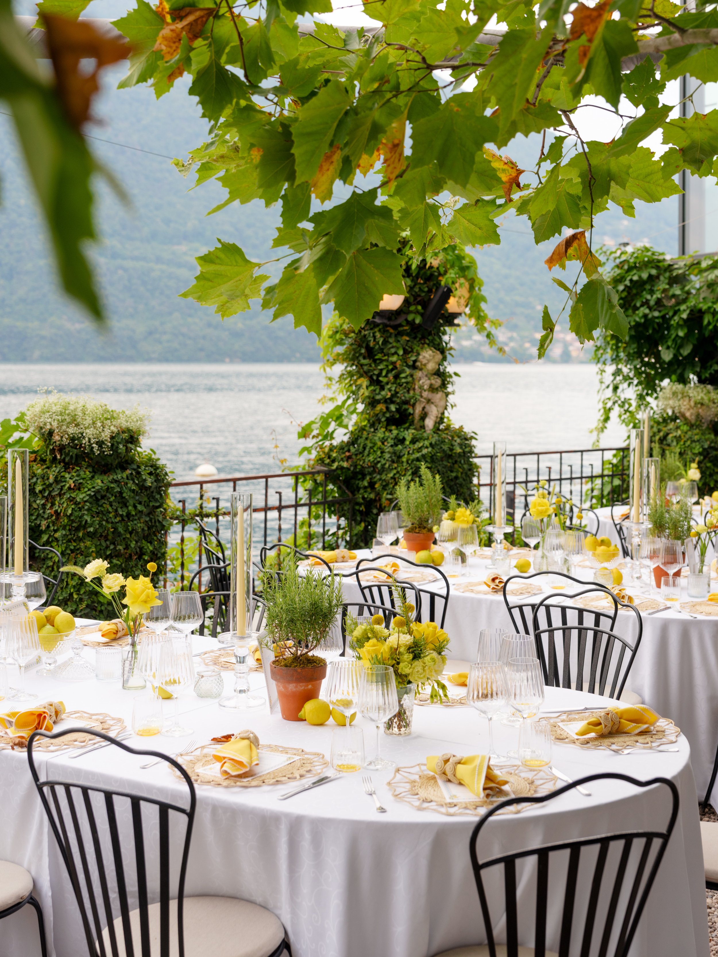 Outdoor dining setup with round tables covered in white tablecloths, decorated with yellow and green floral arrangements, potted herbs, and apples, overlooking a lake with treelined mountains in the background.