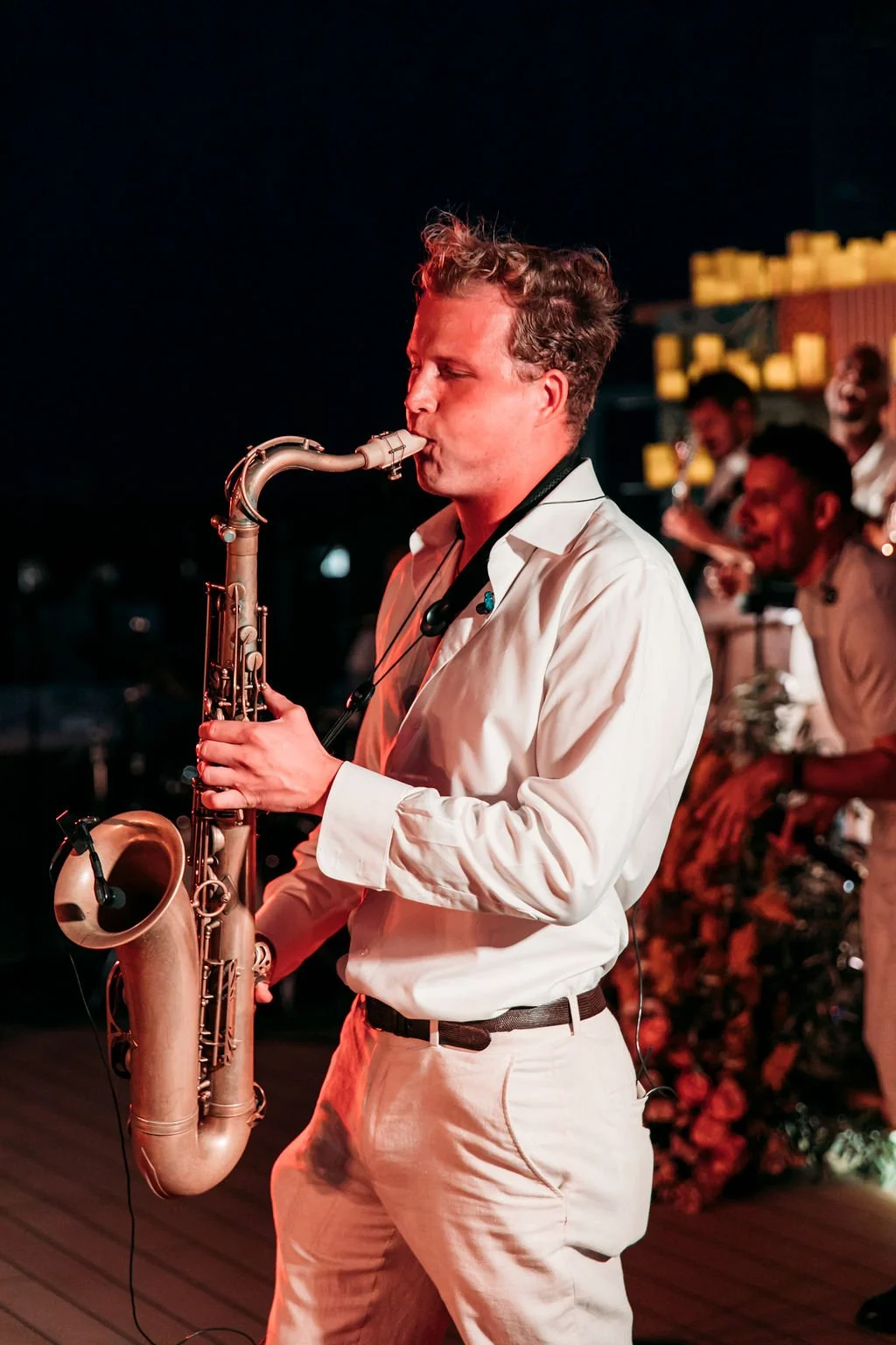 A man in a white shirt and pants playing a saxophone during a nighttime music performance, with several people playing instruments in the background.