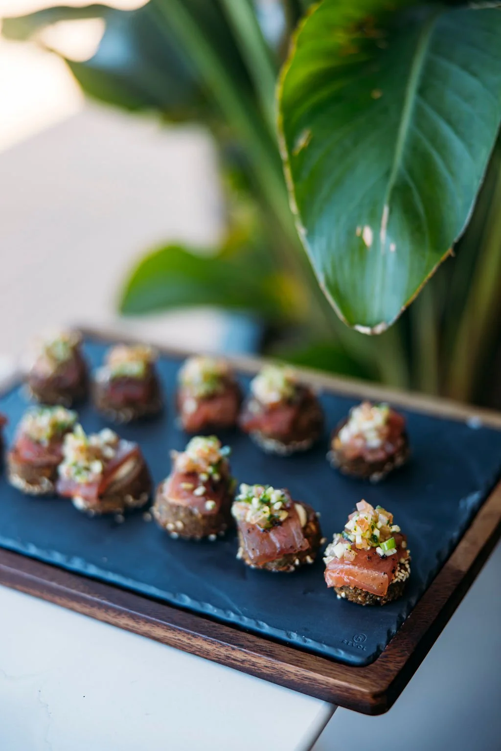 A tray of assorted appetizers or small bites garnished with chopped vegetables, set on a black slate serving board with a large green leafy plant in the background.
