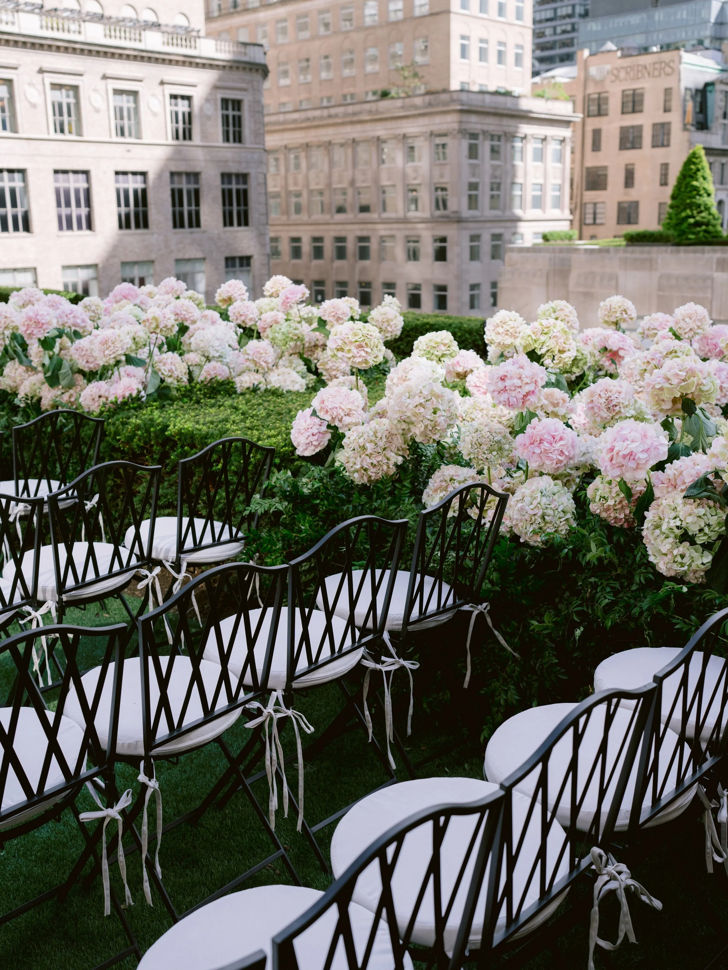 Black chairs with white cushions arranged on a green lawn, with pink and white hydrangea flowers in the background, set on a rooftop garden with city buildings in the distance.