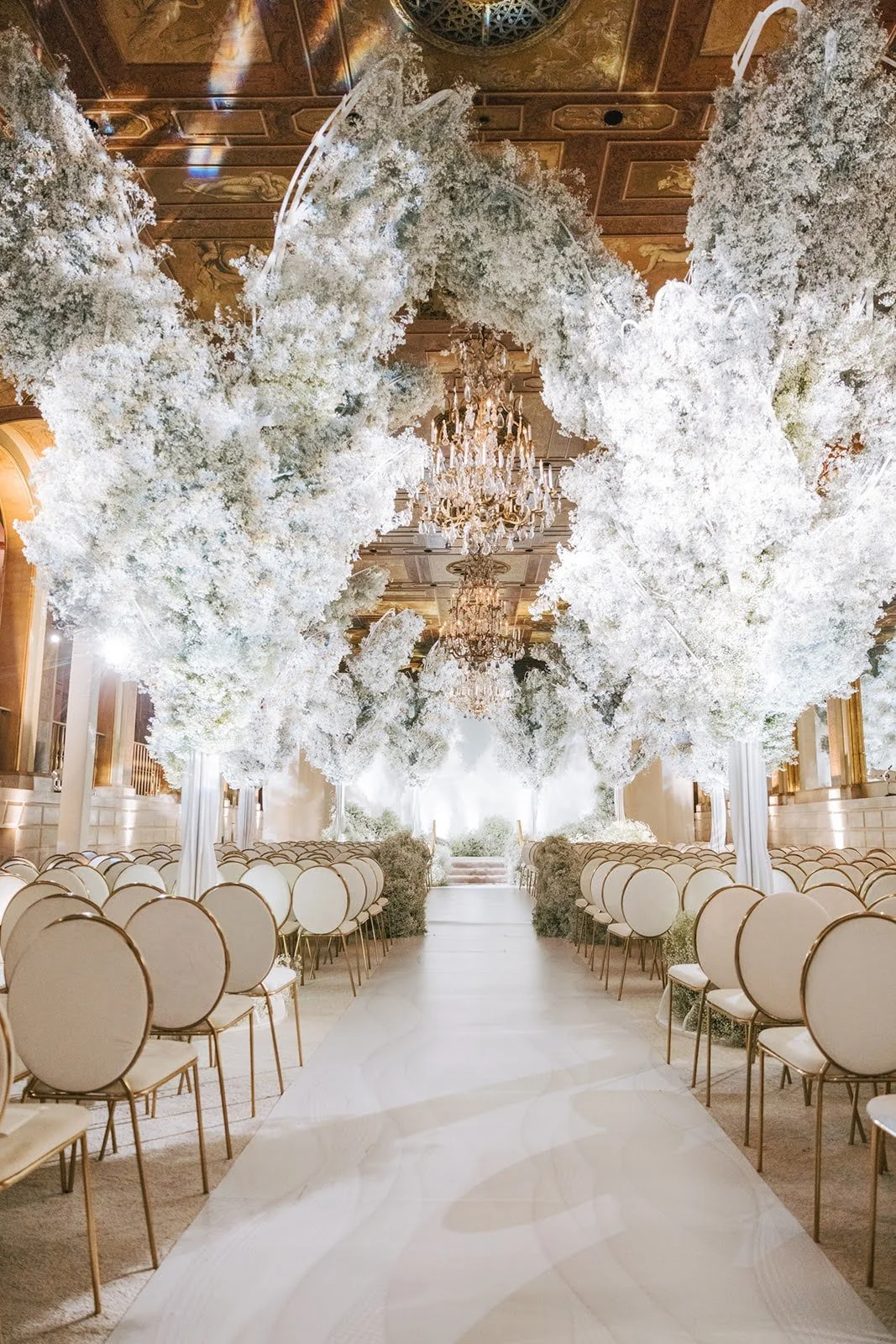 Inside a grand wedding venue with a white aisle, large white floral arrangements hanging from the ceiling, and chandeliers.