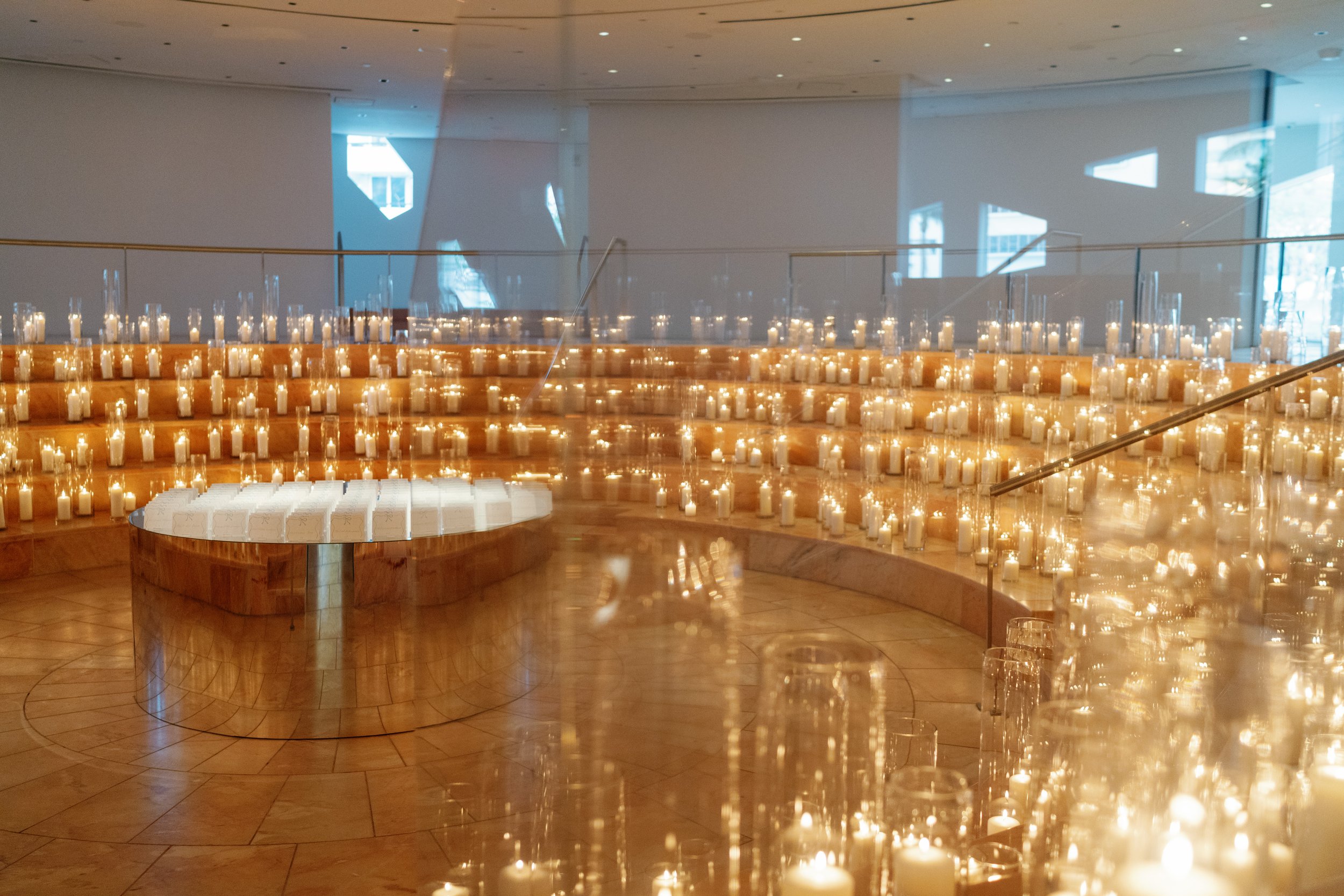 Interior of a modern venue with multiple lit candles arranged on tiered steps and a round table, reflecting candlelight with windows and natural light in the background.