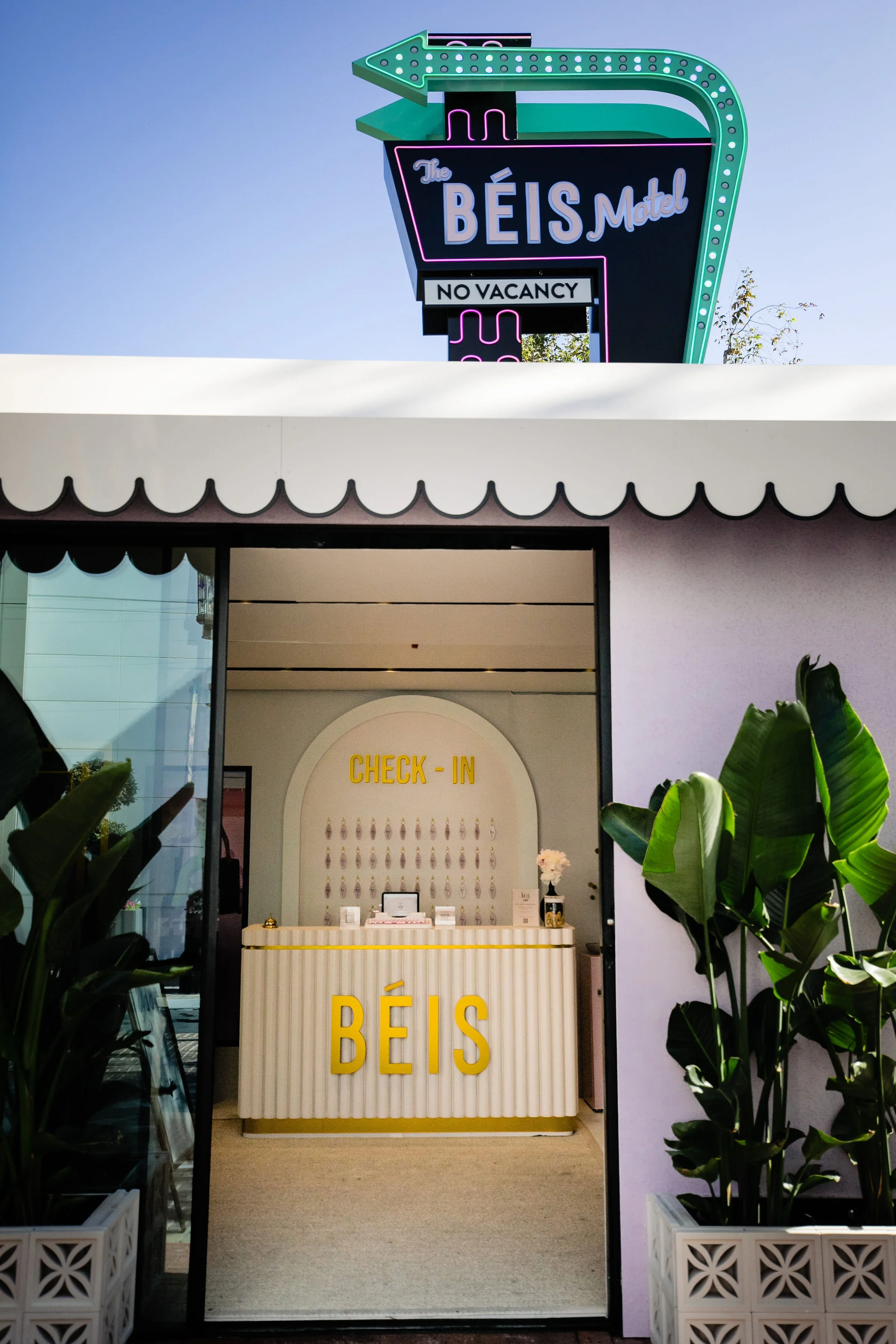 Photo of the entrance to a boutique with a check-in desk labeled "BÉIS" and a neon sign above indicating the BEIS Motel with a "No Vacancy" sign, surrounded by potted plants.