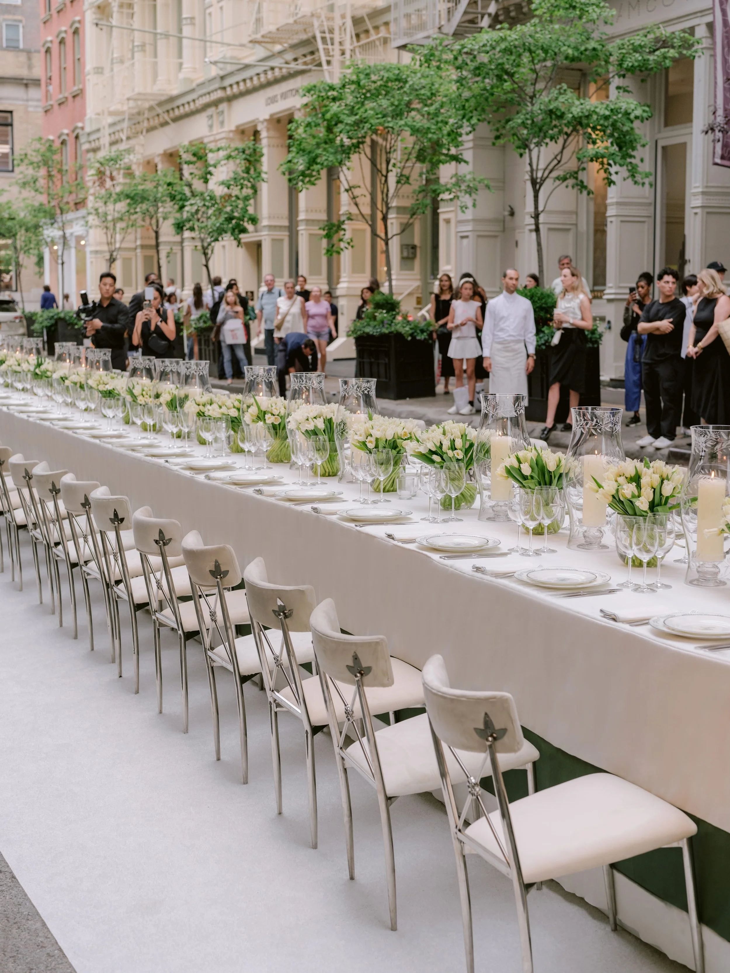 Long wedding reception table set with white flowers and glassware, outside on a city street with people and trees
