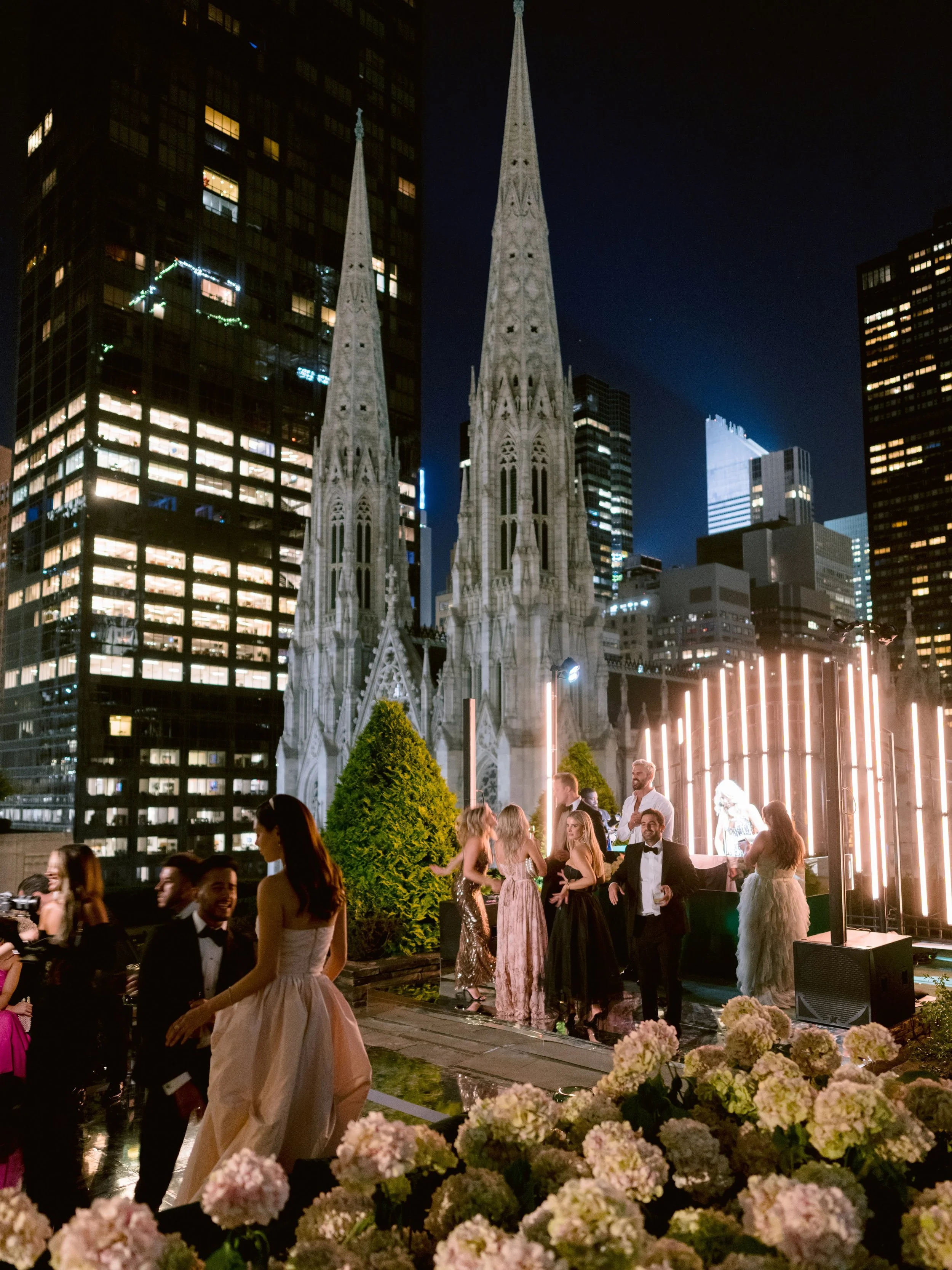 Nighttime rooftop party in New York City with guests dancing and socializing, a church with twin spires in the background, illuminated modern buildings, and flowers in the foreground.