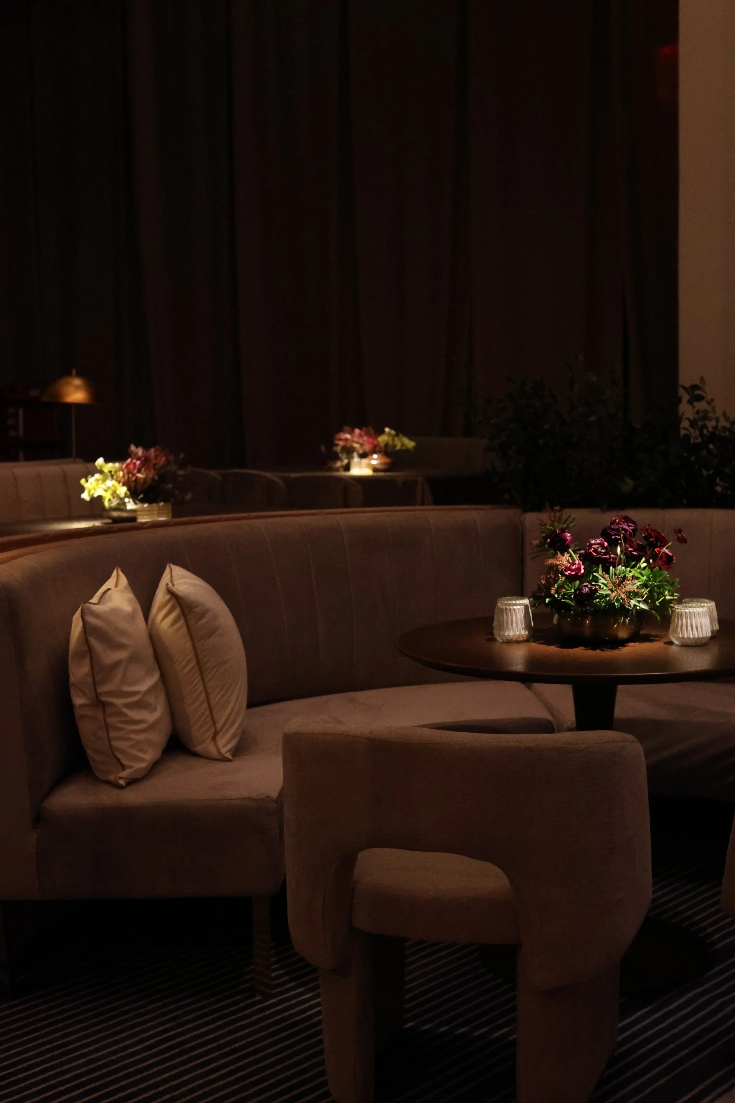 A dimly lit lounge with round tables decorated with flower arrangements and candles, plush beige seating, and dark curtains in the background.