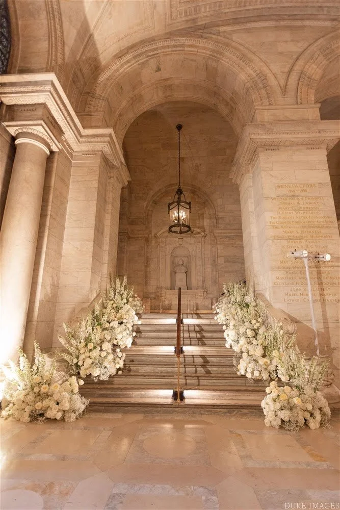 Elegant indoor staircase decorated with white and cream floral arrangements on both sides, with a stone statue at the top and a vintage hanging lantern above. The setting appears to be a memorial or ceremonial hall.