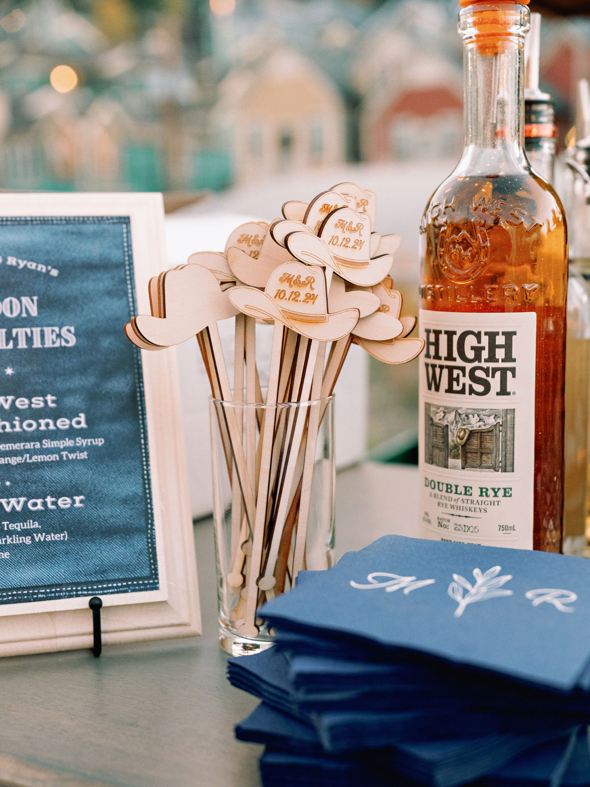 A metal surface with a blue napkin that has white cursive writing on it, a tall glass filled with wooden cocktail stir sticks, and a bottle of High West Double Rye whiskey. The stir sticks have cowboy hat designs with the date 10.12.21 printed on the