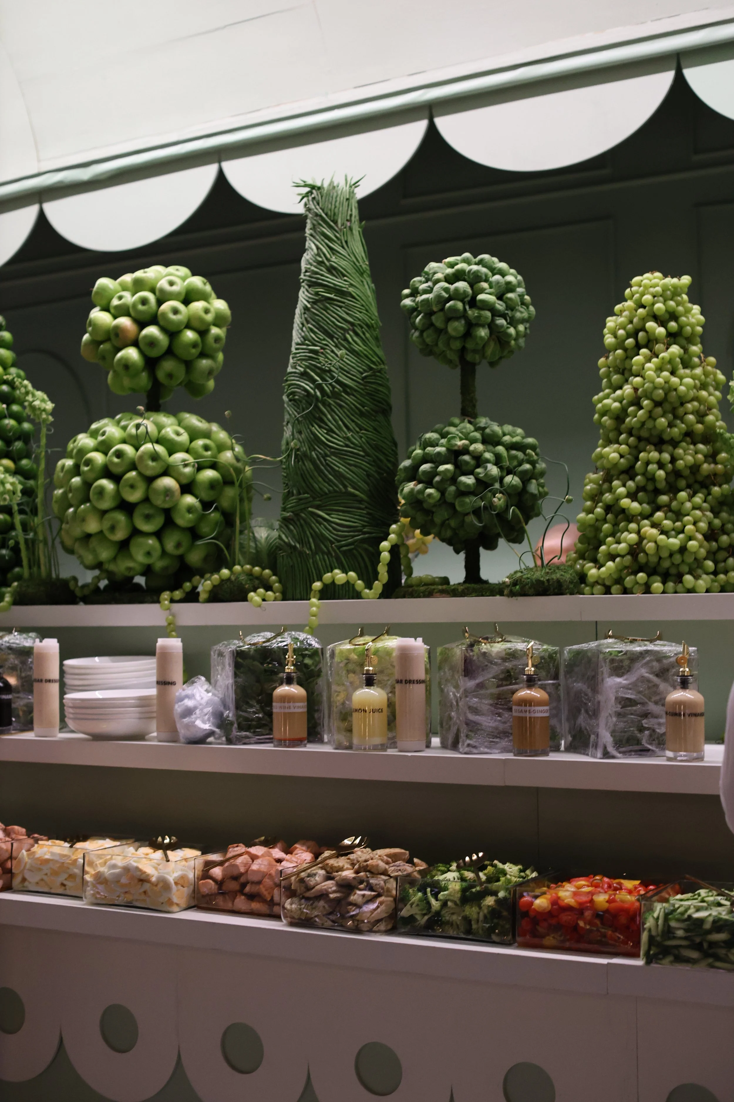 Display of green decorative trees made of apples and other fruits, with bottles and bowls in front of them at a buffet or food display.