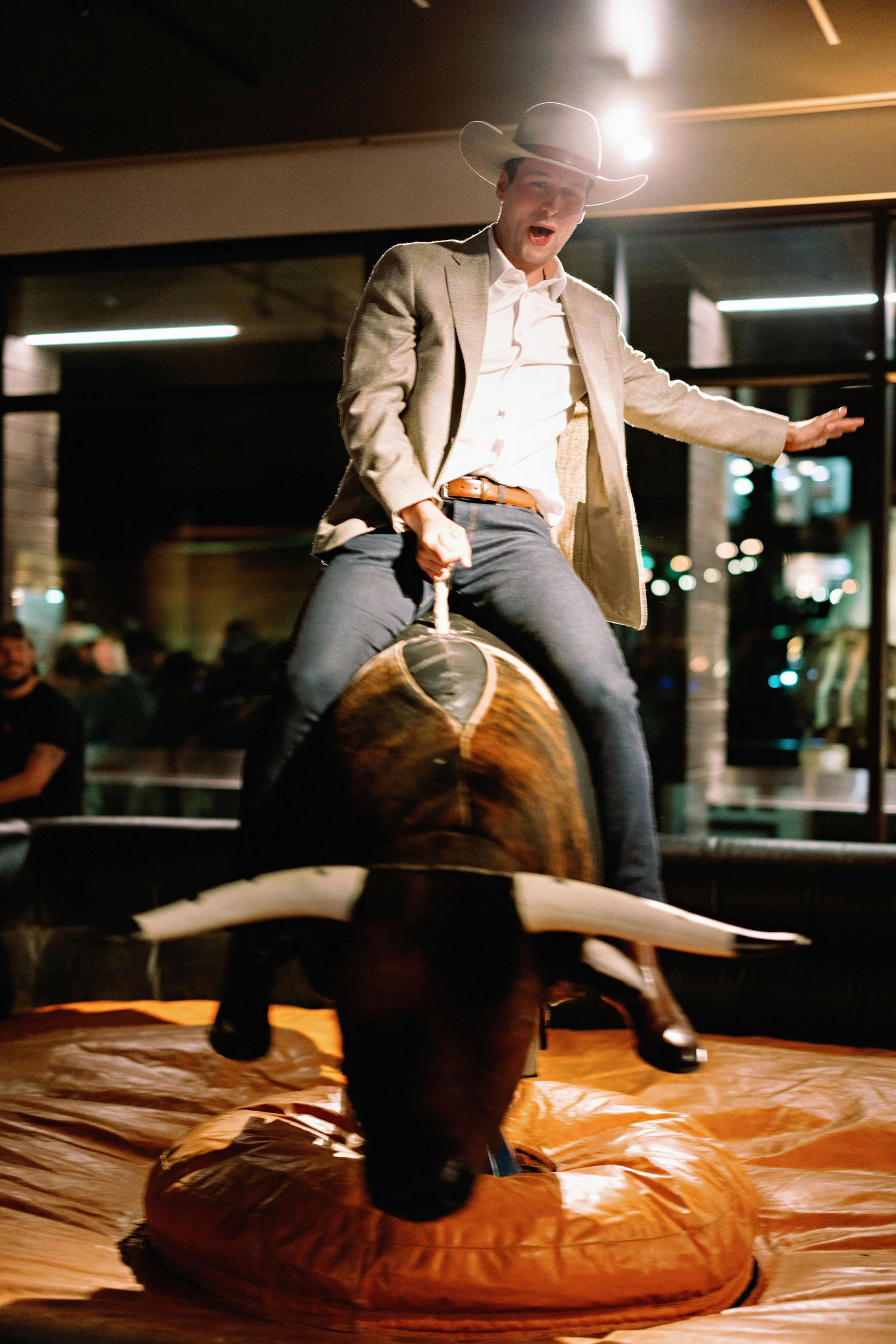 A young man in a cowboy hat and suit jacket riding a mechanical bull in a dimly lit indoor setting with people in the background.
