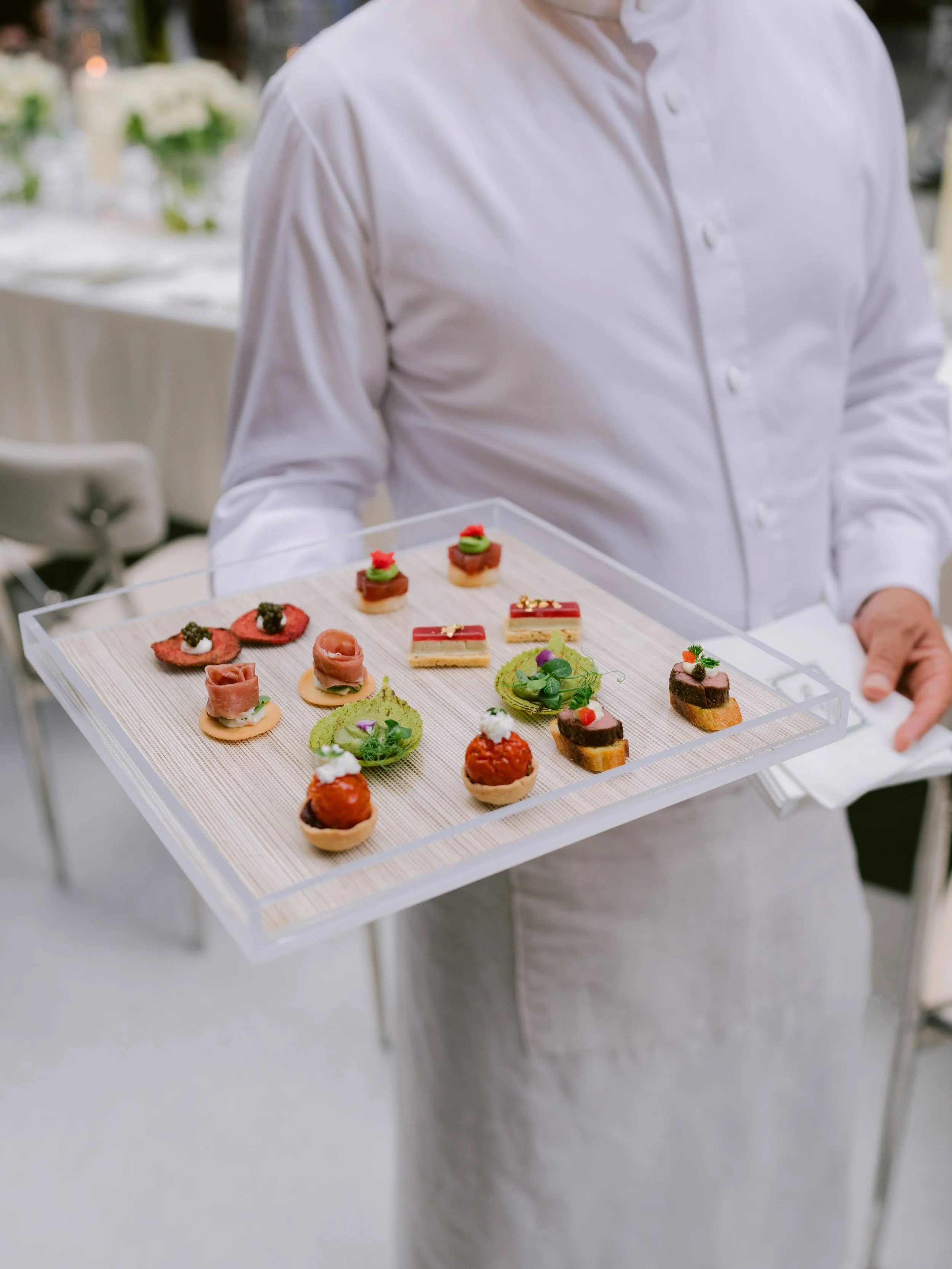 Person in white shirt and apron holding a tray of assorted small, decorative appetizers and desserts at a catering event.