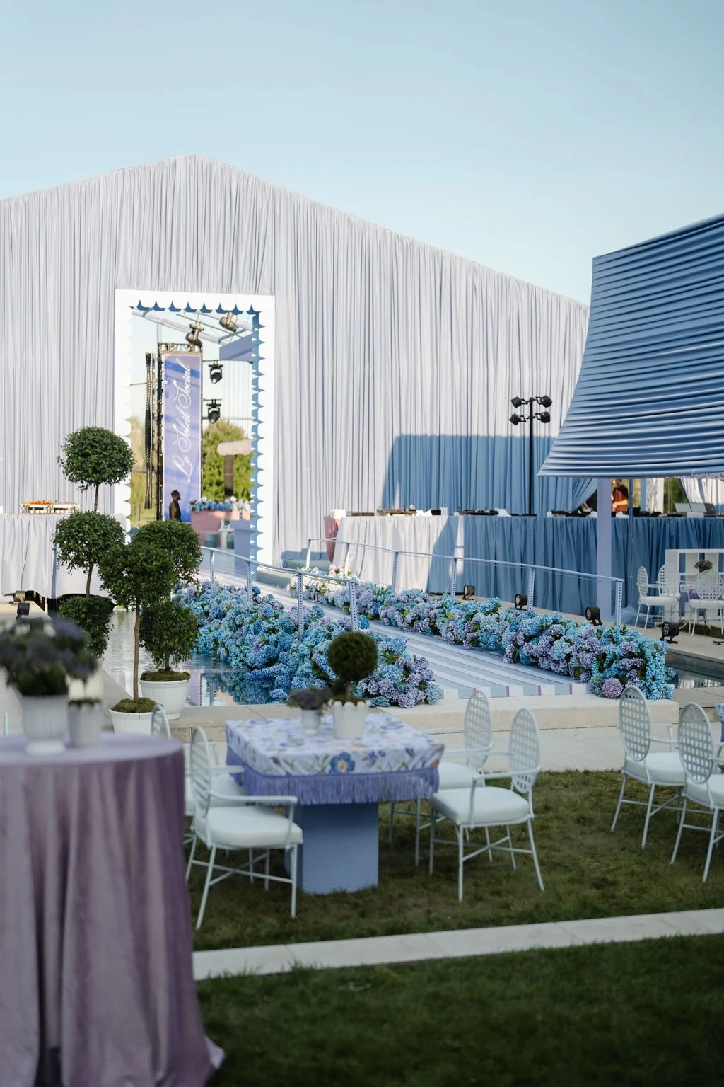 Outdoor event setup with decorated tables, flowering plants, and a stage with a white draped backdrop and a large mirror frame, under clear blue sky.