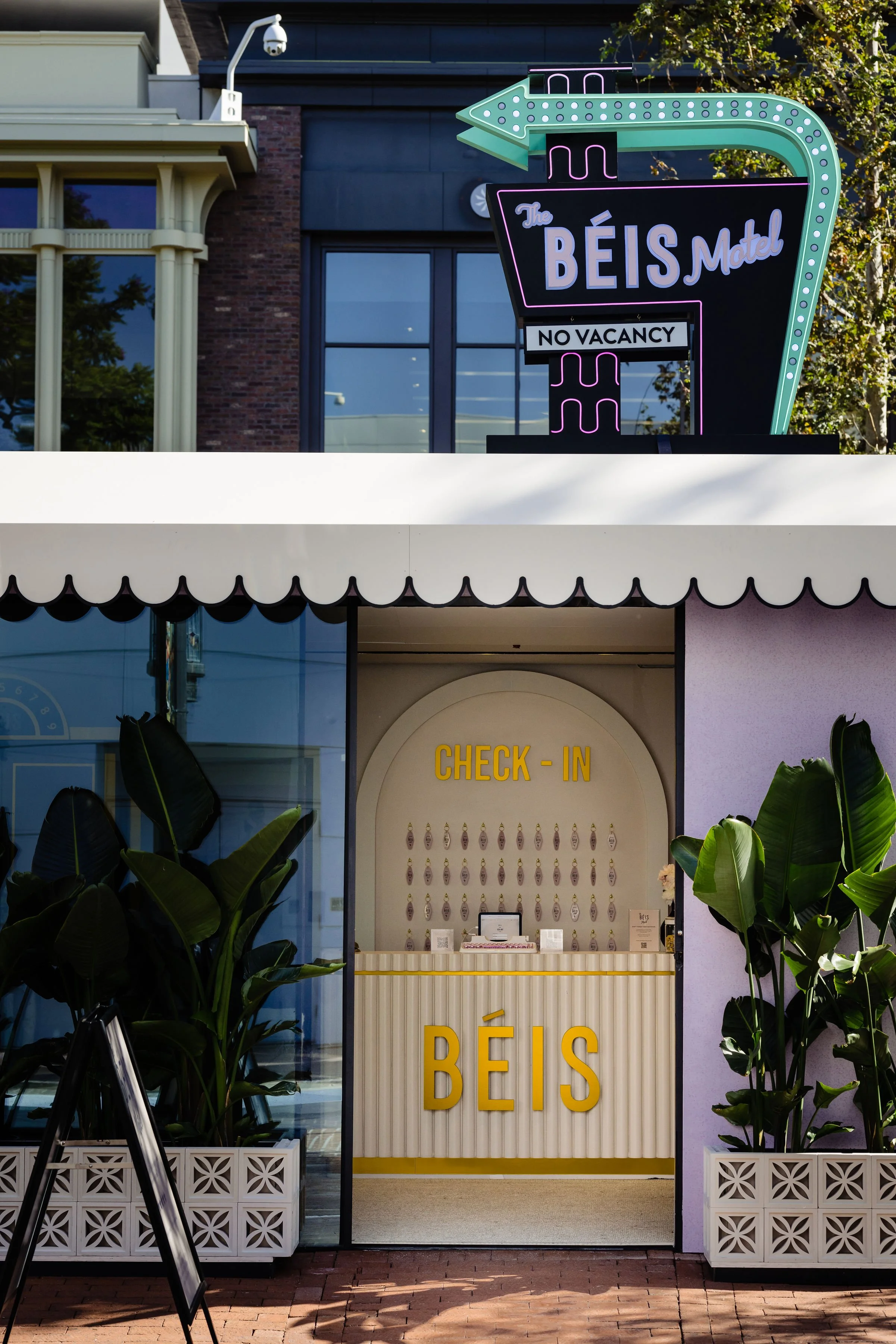 Sign for The BÉIS Motel with neon ice cream cone design, indicating no vacancy. Below, the check-in counter for BÉIS with a white and yellow theme, surrounded by green plants.