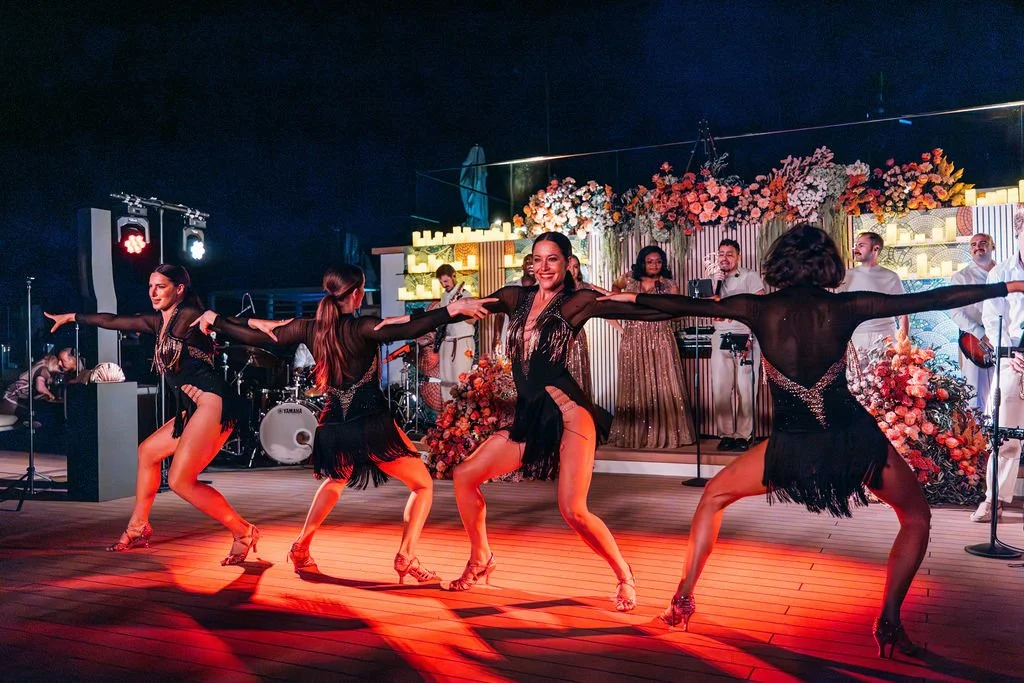 Five women in dance costumes performing a choreographed routine on a stage decorated with flowers and backlit at night. A band is playing in the background.