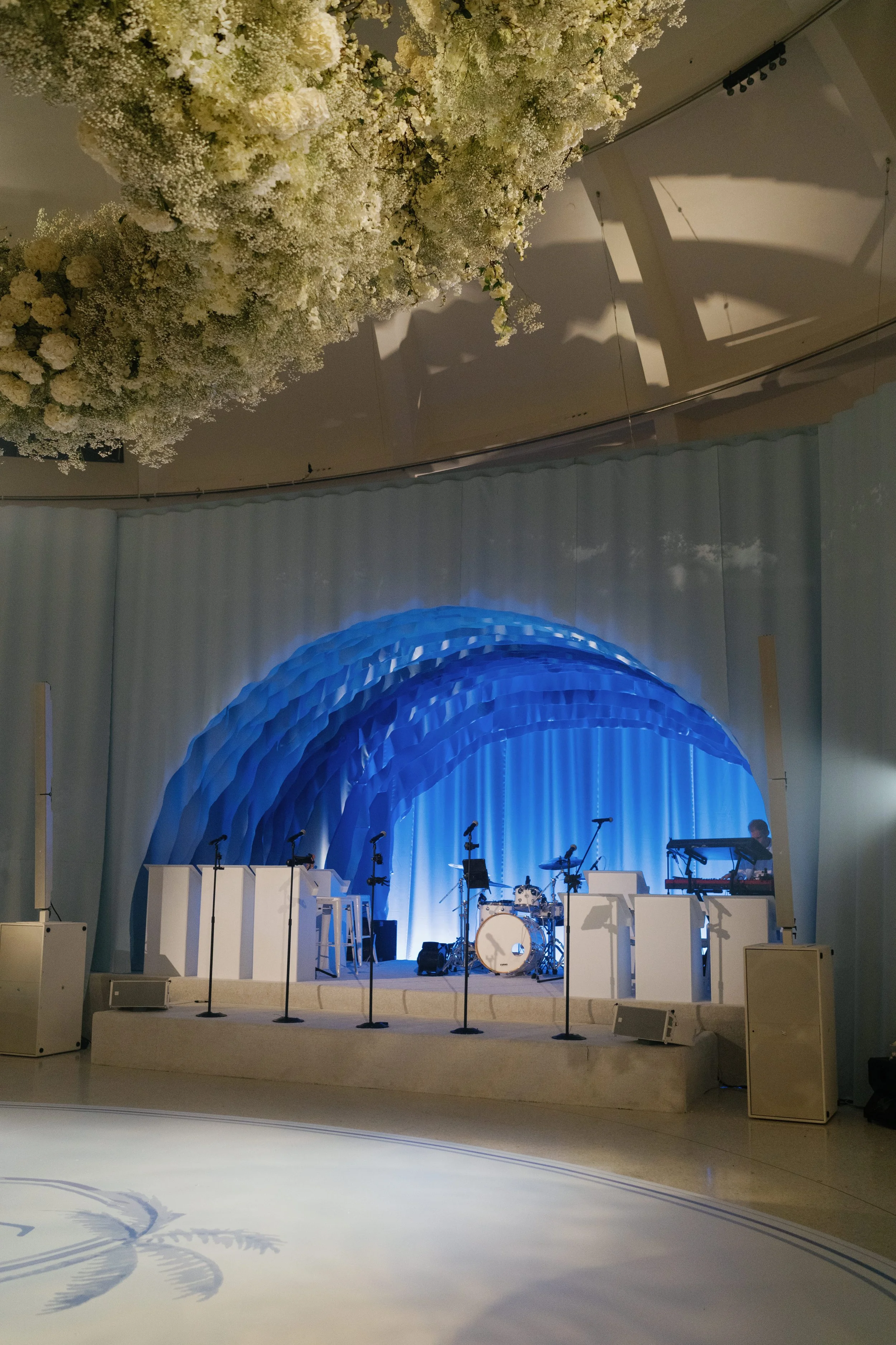 Stage setup with musical instruments including drums, keyboards, and microphones, decorated with a blue arch and white seating, in front of a light blue curtain, with floral arrangements hanging from the ceiling.