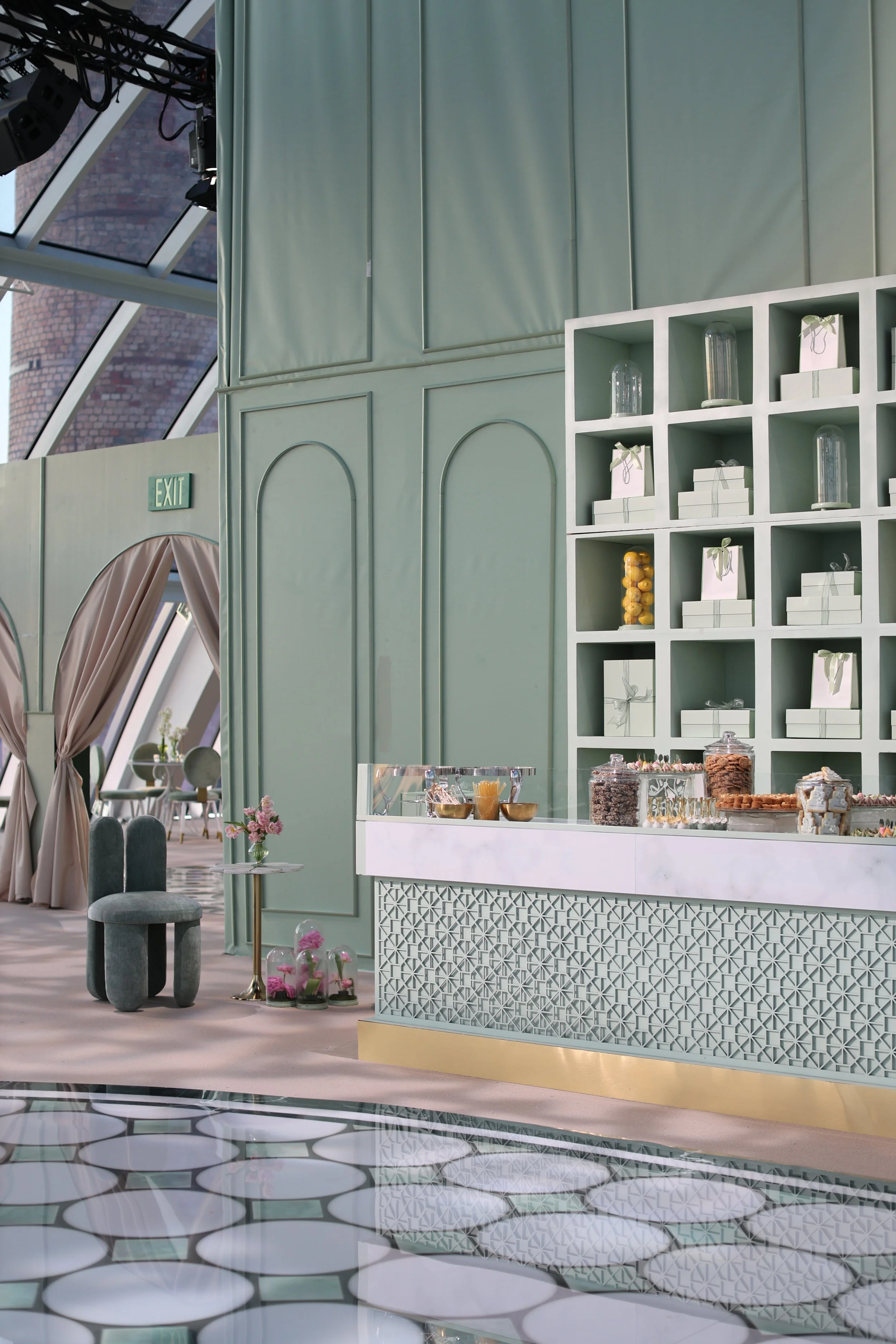 Interior of a pastel green-themed cafe or event space with a modern white counter displaying sweets and snacks, a white shelving unit with decorative items, and a seating area with velvet chairs and a flower arrangement, illuminated by natural light 