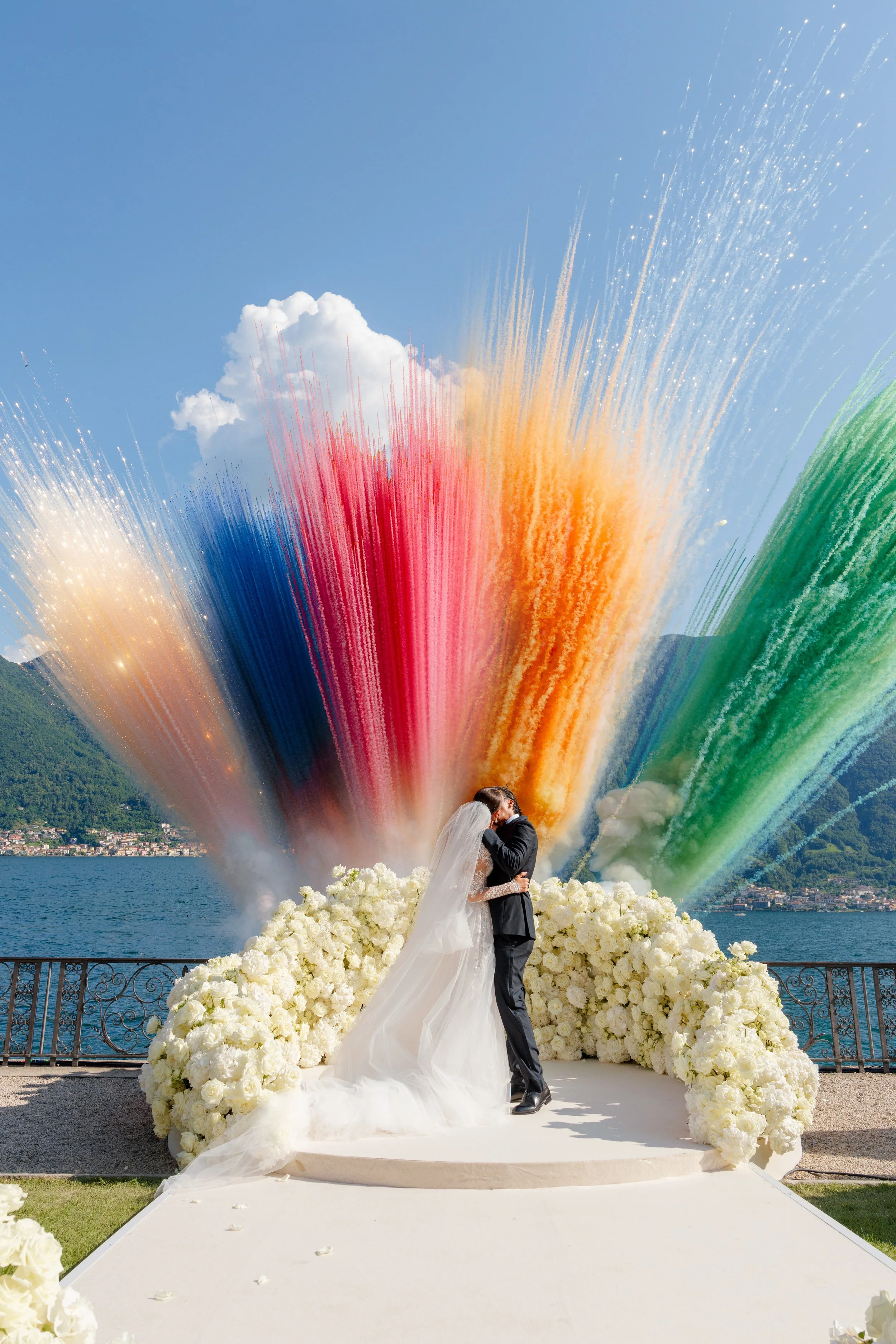 A bride and groom kissing on a wedding altar decorated with white flowers, with colorful fireworks in the sky above them, near a body of water with mountains in the background.