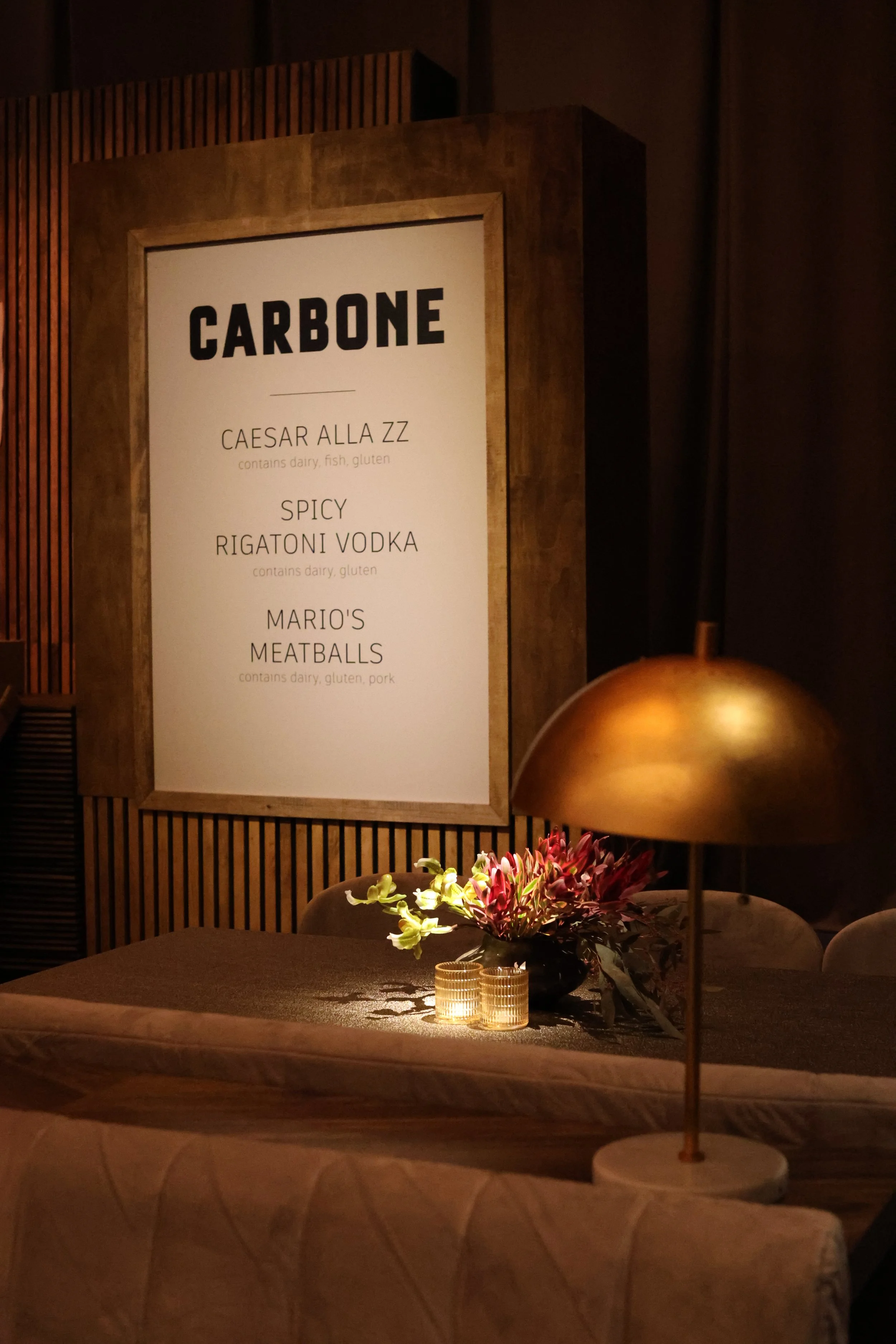 A menu board at a restaurant named Carbone, listing dishes such as Caesar Alla ZZ, Spicy Rigatoni Vodka, and Mario's Meatballs, with a table decorated with flowers and a gold-colored lamp in the foreground.