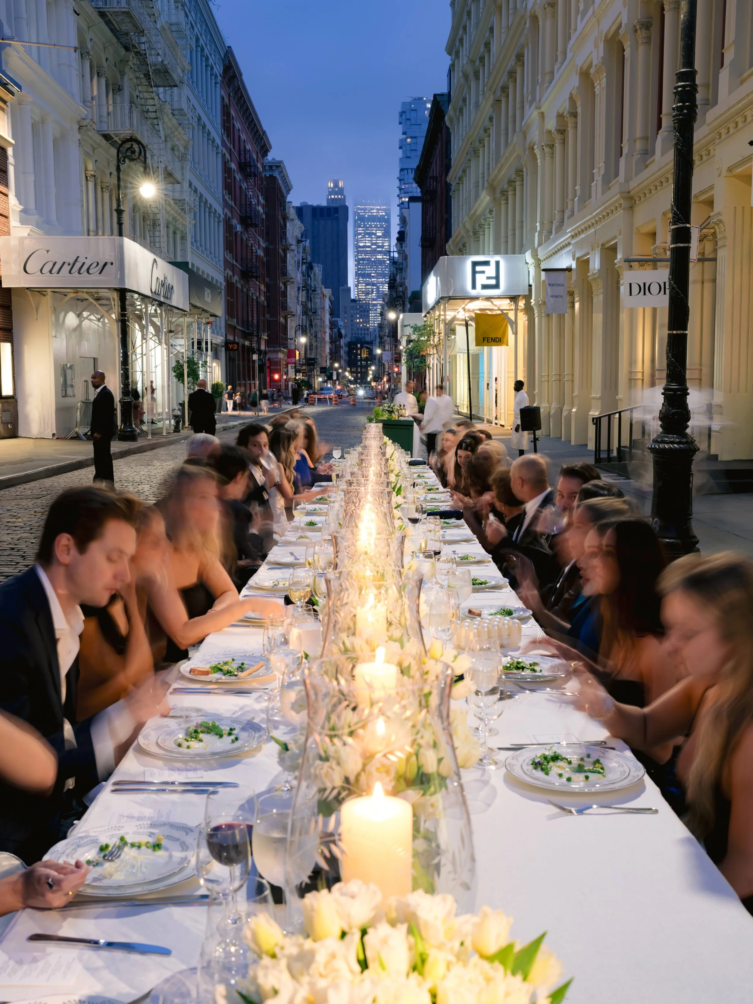 Long dinner table set with candles and floral arrangements on a city street during evening, with guests dining and luxury storefronts in the background.