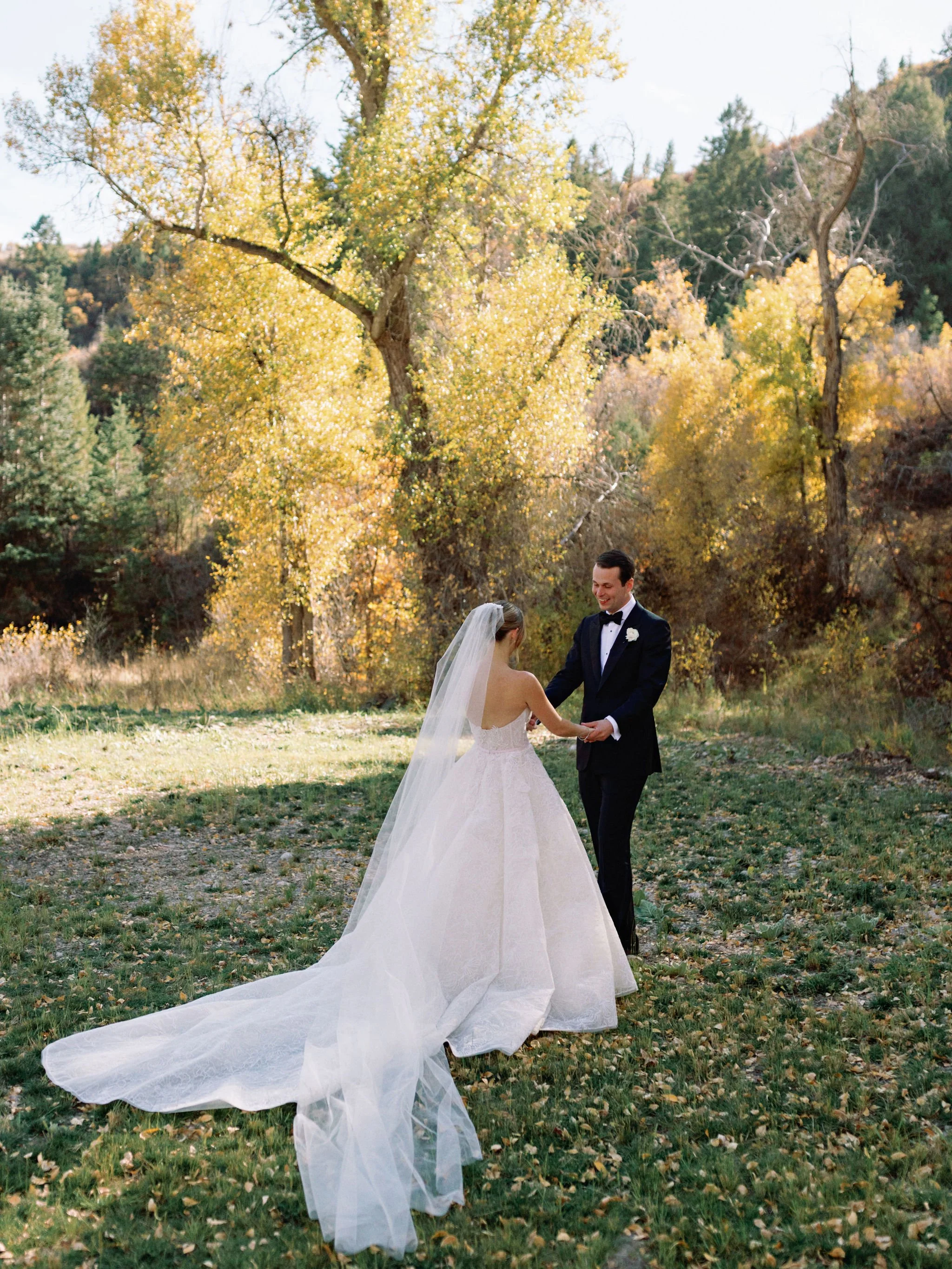A bride and groom holding hands outdoors in a park surrounded by trees with yellow and green leaves during autumn.