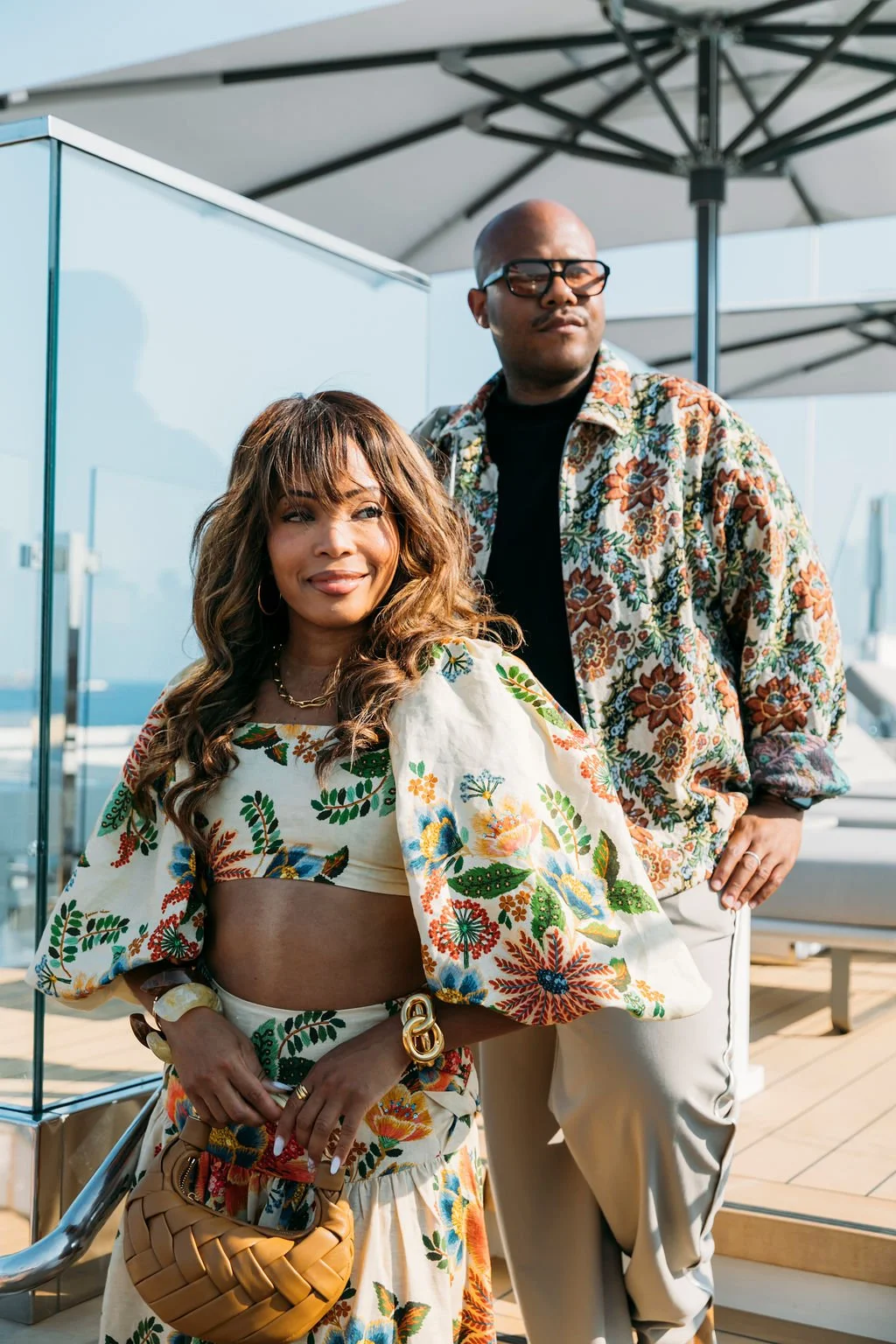 A woman and a man stand outdoors on a balcony or terrace, dressed in colorful, floral-patterned clothing. The woman is smiling and holding a tan woven handbag, while the man stands behind her with his hand on his hip, looking off into the distance.