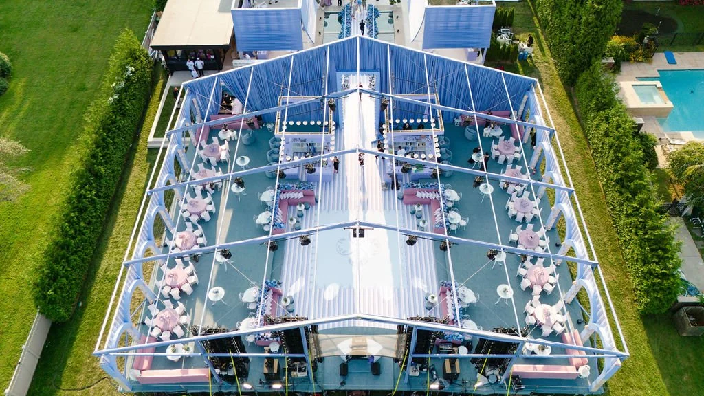 An aerial view of an event tent set up outdoors with pink and white decorations, tables with pink tablecloths, and chairs, surrounded by green lawns and a swimming pool nearby.