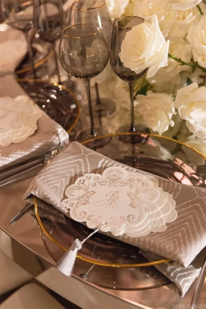 Elegant table setting featuring a gold-rimmed glass plate with a intricately designed beige napkin and a decorative place card. In the background, there are two black wine glasses and a floral arrangement with white roses.