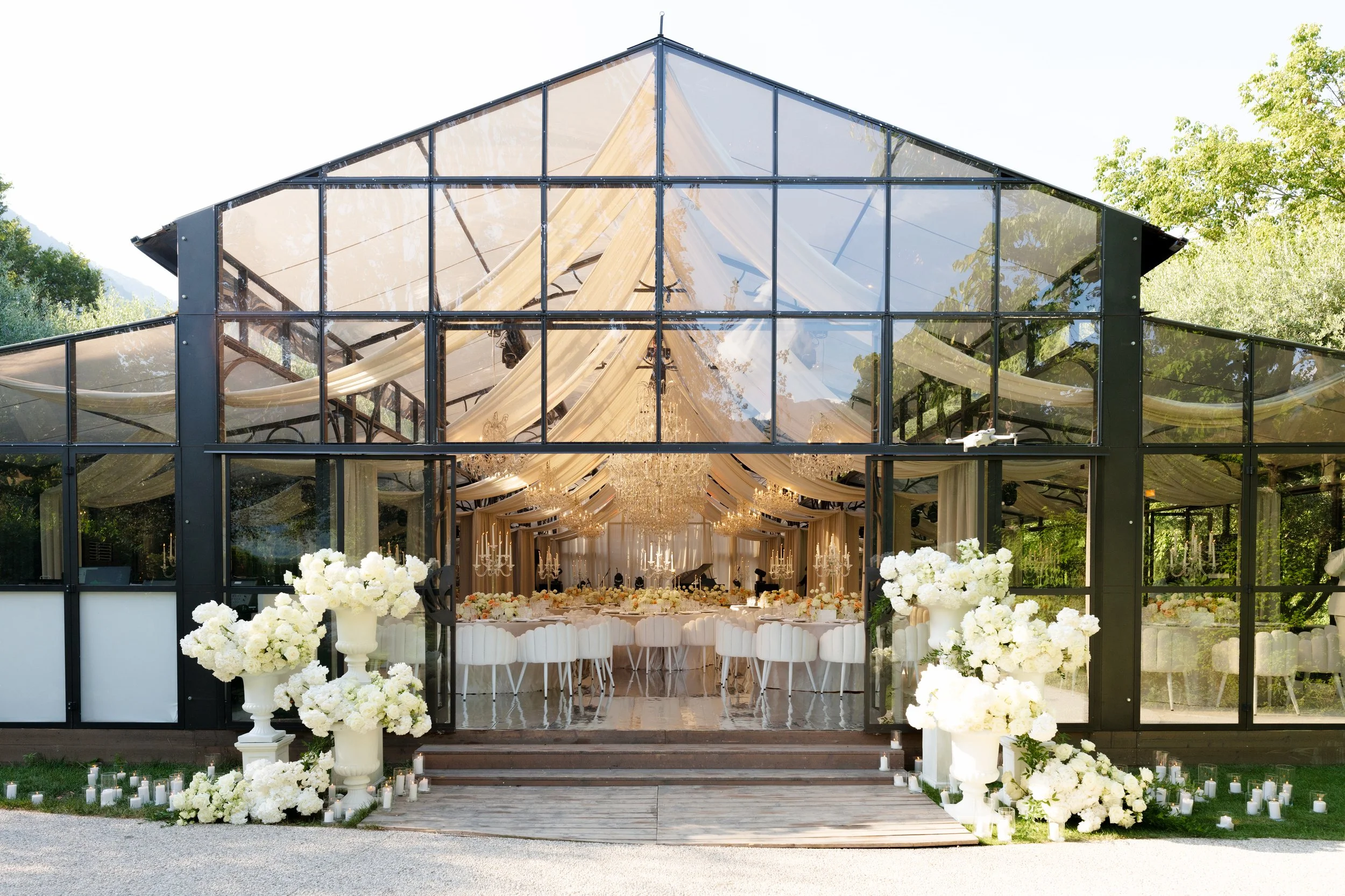 A glass-walled event venue decorated in white for a wedding reception to be held outdoors, with floral arrangements and candles at the entrance and elegant drapery inside.