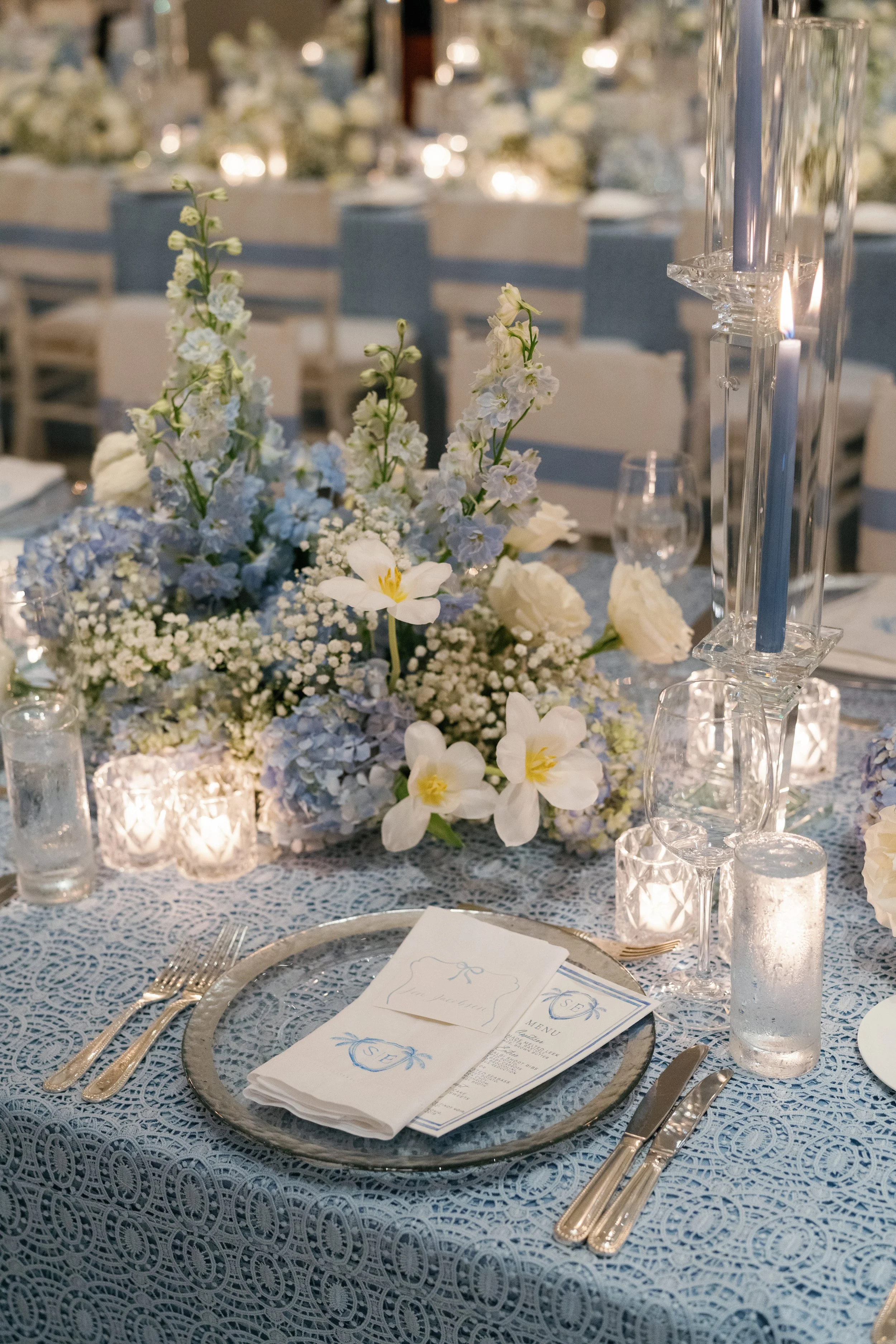 Elegant wedding reception table with blue and white floral centerpiece, candles, and place settings on a lace tablecloth.