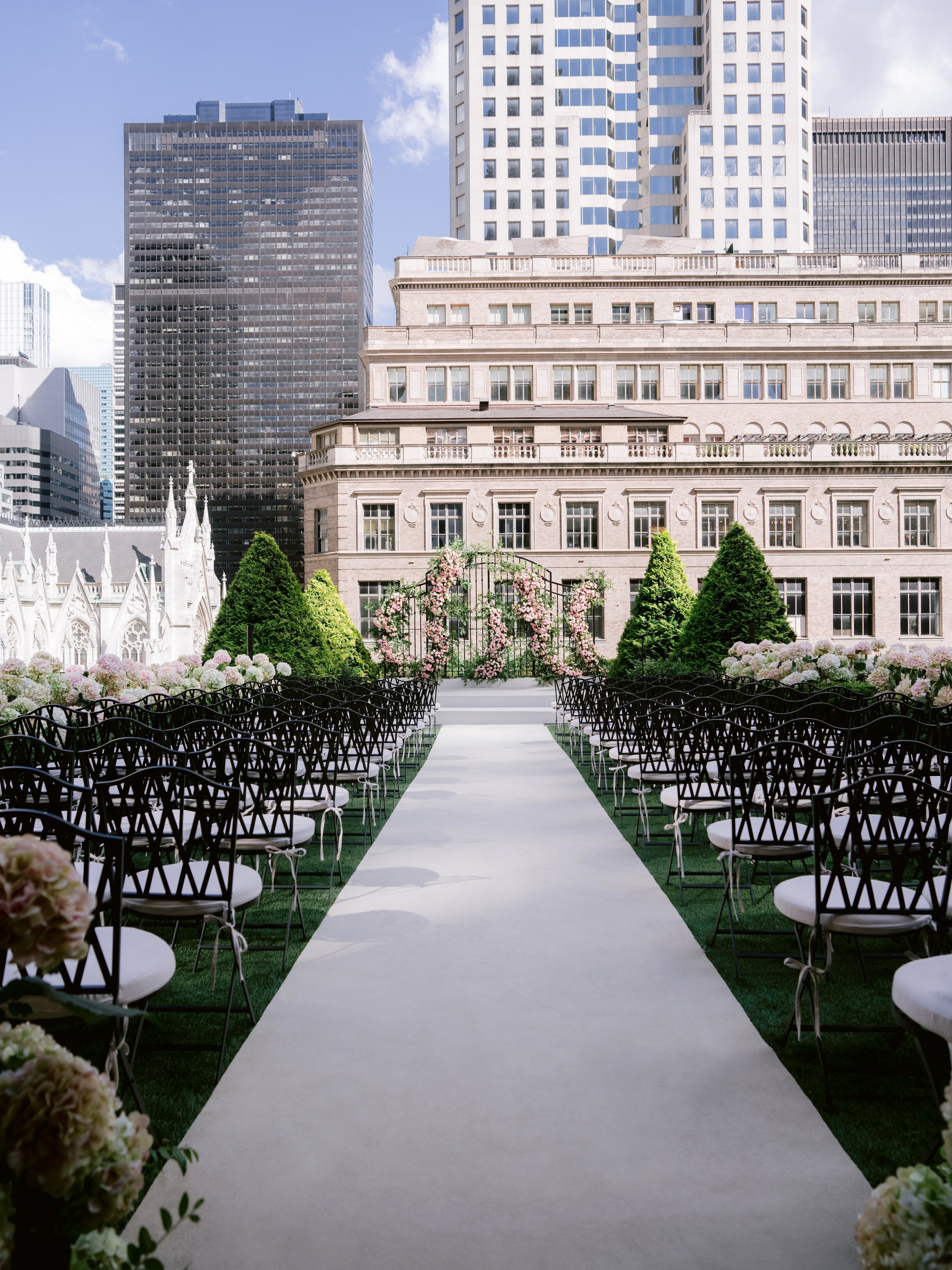 Outdoor wedding ceremony setup with chairs arranged on either side of a white aisle, floral decorations, and green shrubbery in front of tall buildings in an urban setting.