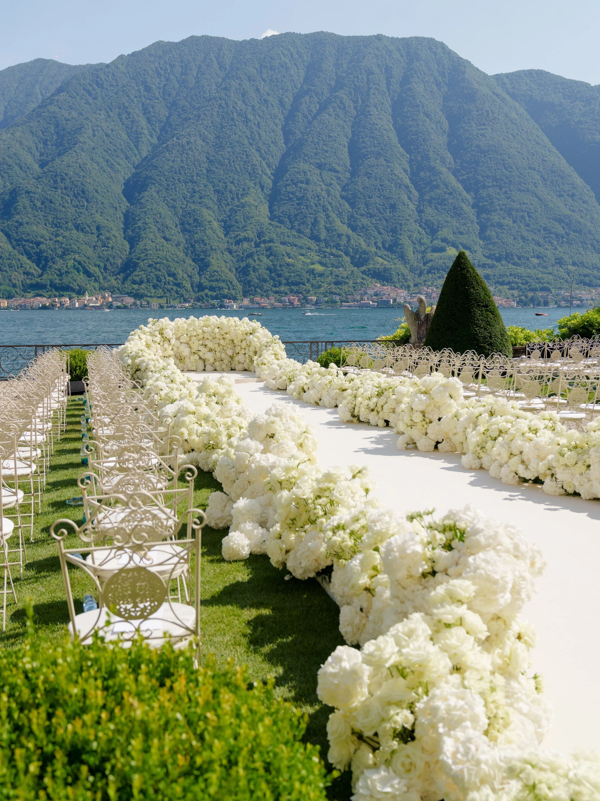 Outdoor wedding ceremony setup with white floral aisle, chairs, and scenic mountain background.