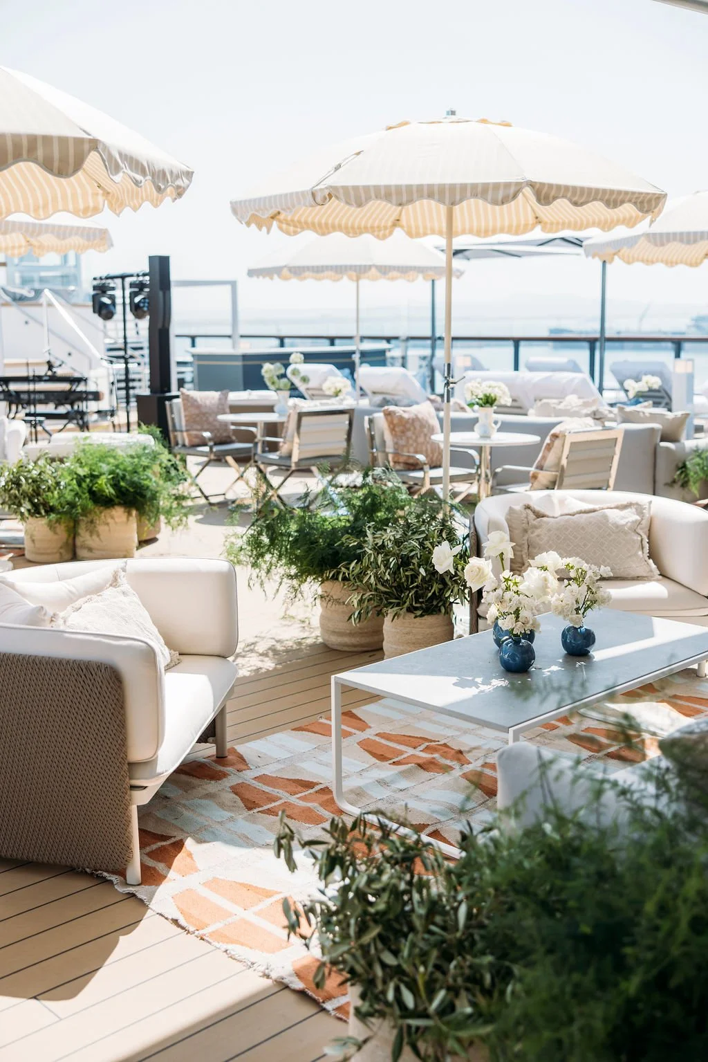 A sunny outdoor rooftop lounge area with white sofas, glass vases with white flowers, umbrellas, potted greenery, and a view of the city and ocean in the background.